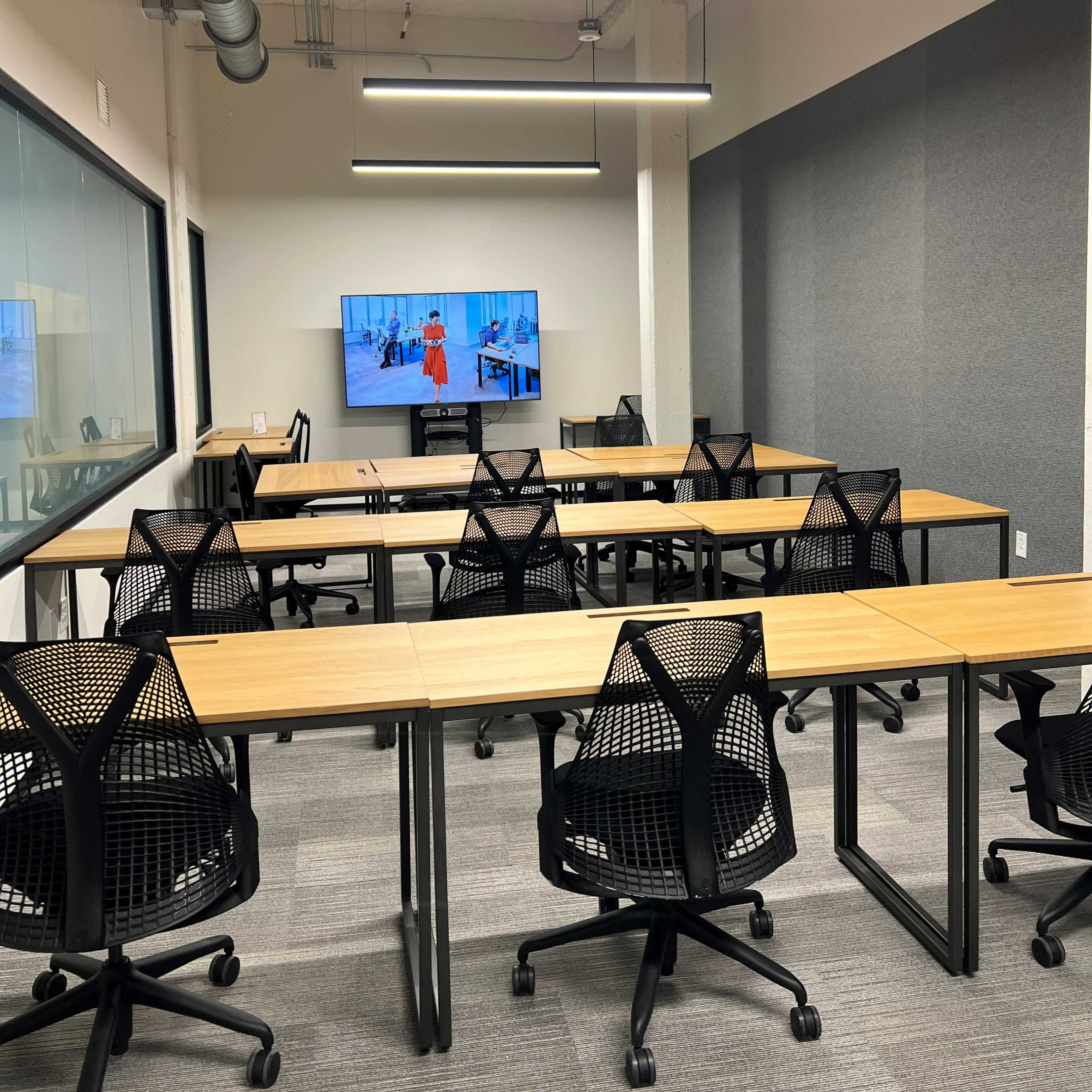 A modern conference room features multiple tables and chairs arranged with a large screen displaying a presentation on one wall.