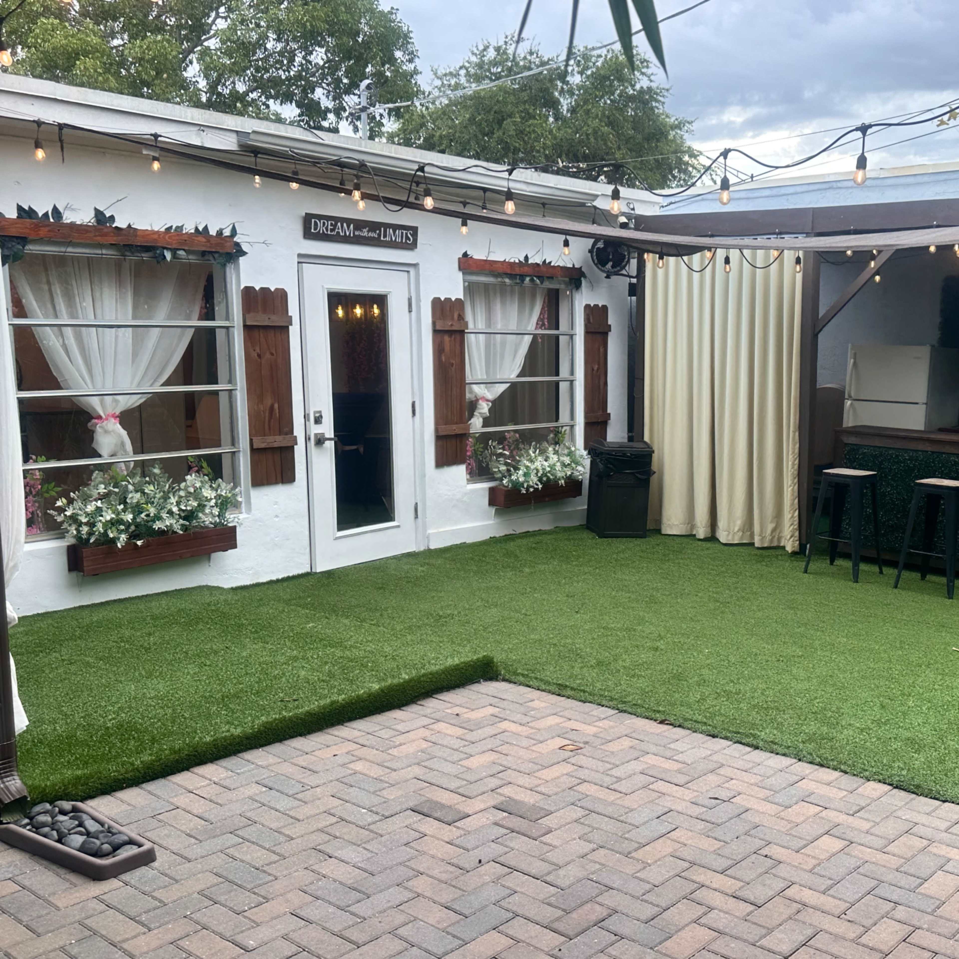 An outdoor space featuring a bar with stools, surrounded by greenery and decorative curtains, on a paved area.