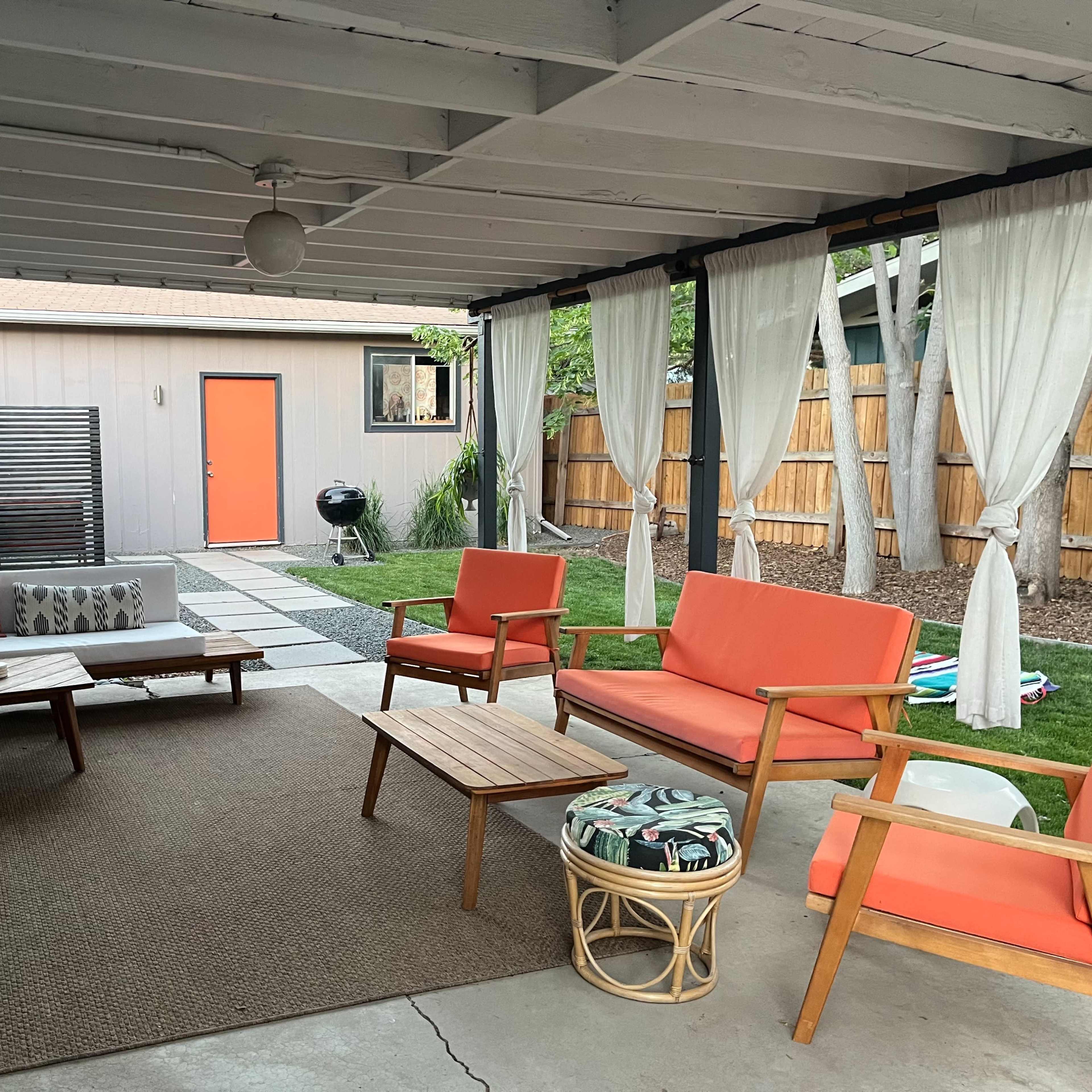 A covered outdoor living space with orange chairs, a wooden table, and light curtains, overlooking a landscaped backyard with grass and stones.