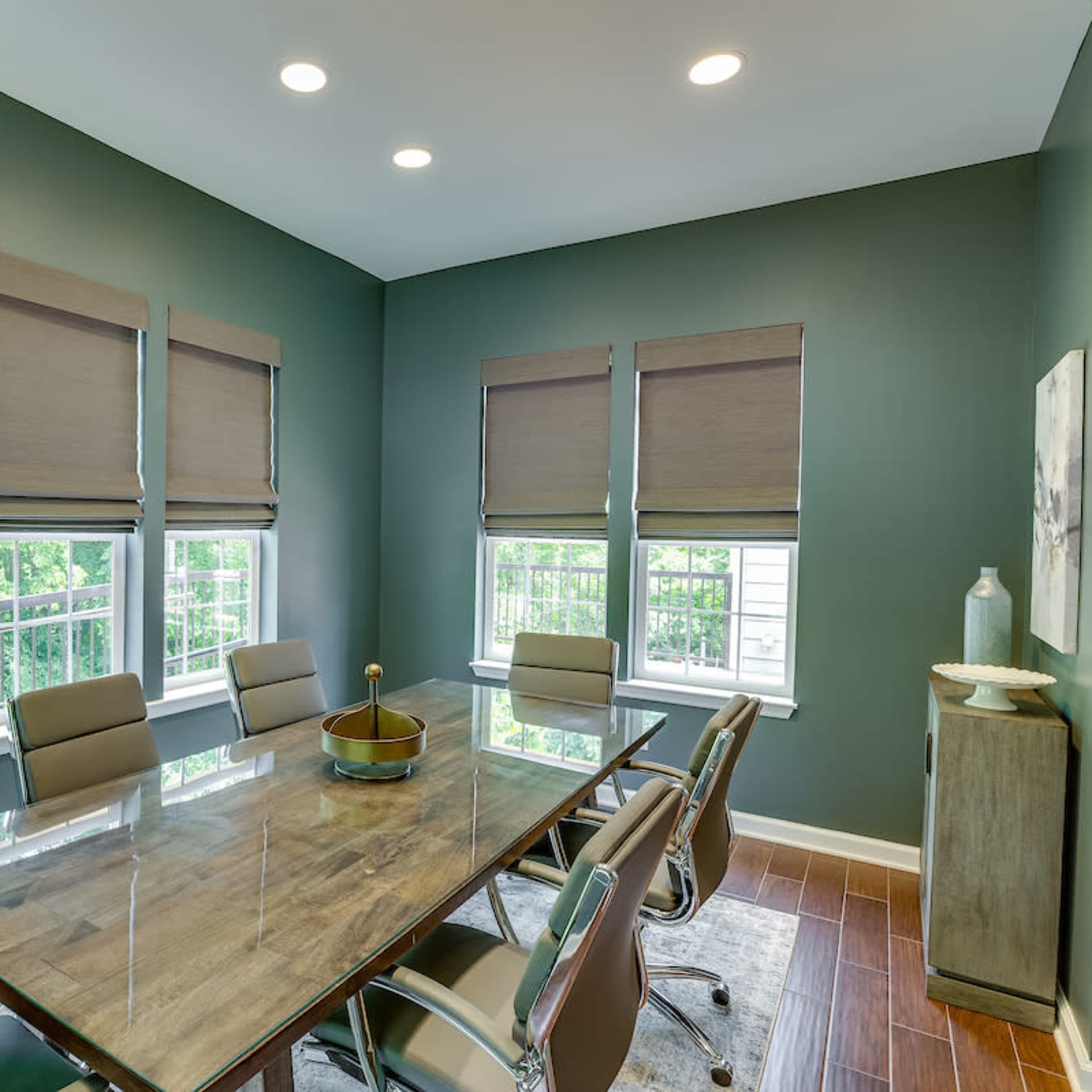 A modern conference room features a rectangular glass table surrounded by ergonomic chairs, with large windows dressed in wooden blinds letting in natural light.