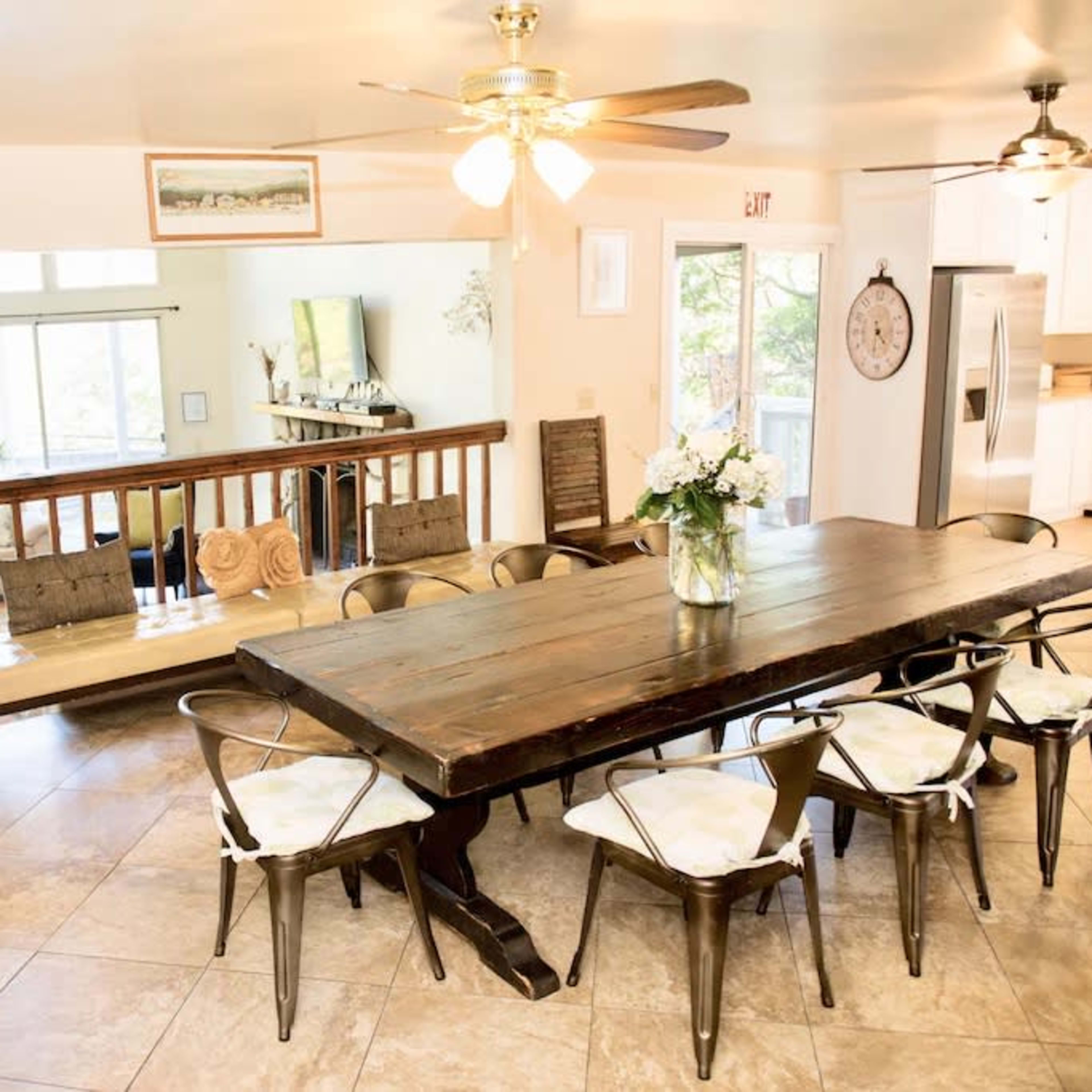 A spacious dining area with a large wooden table surrounded by metal chairs, opening up to a brightly lit living space in the background.