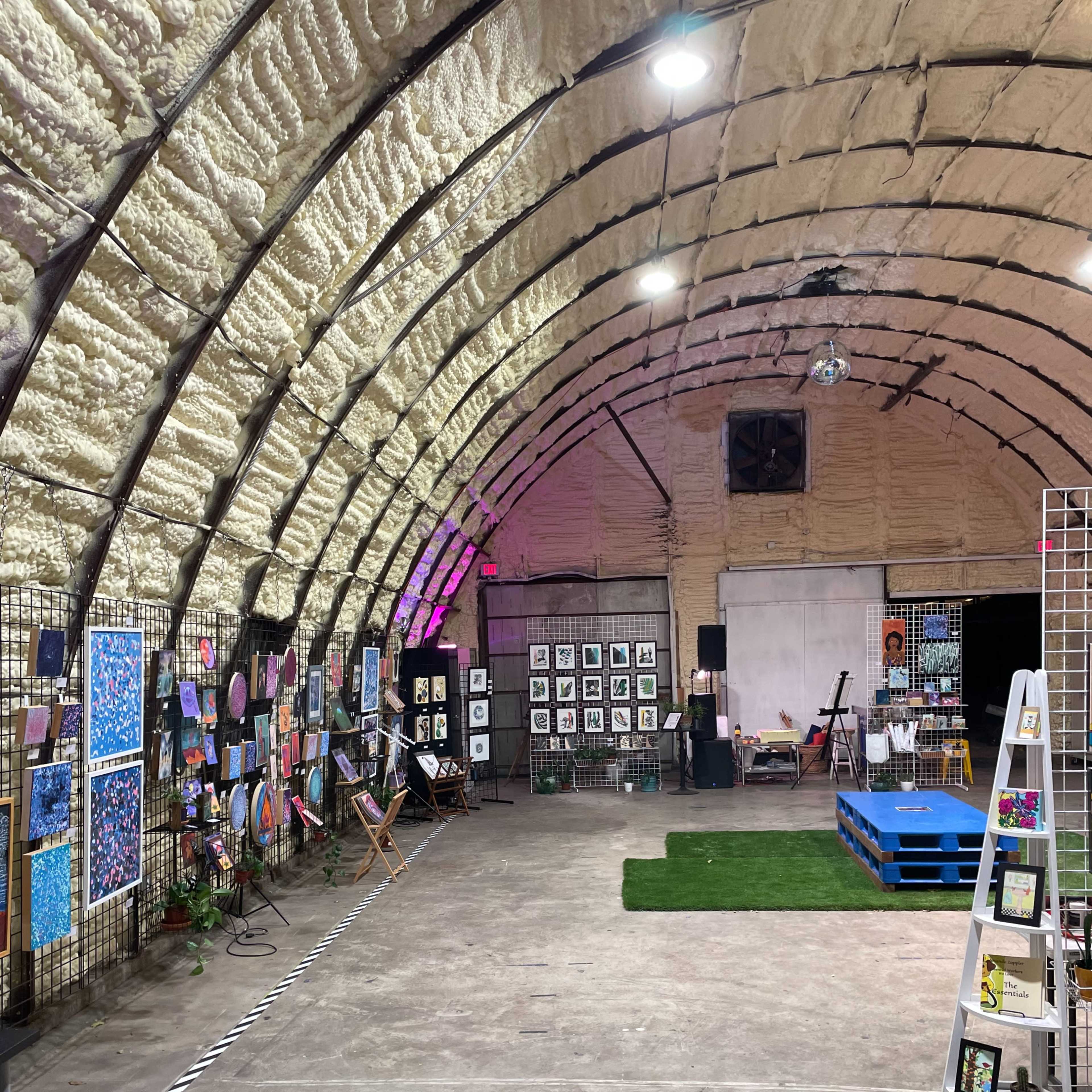 An indoor art exhibit featuring various paintings and artworks displayed on walls with a curved ceiling, set in a spacious, industrial-style environment.