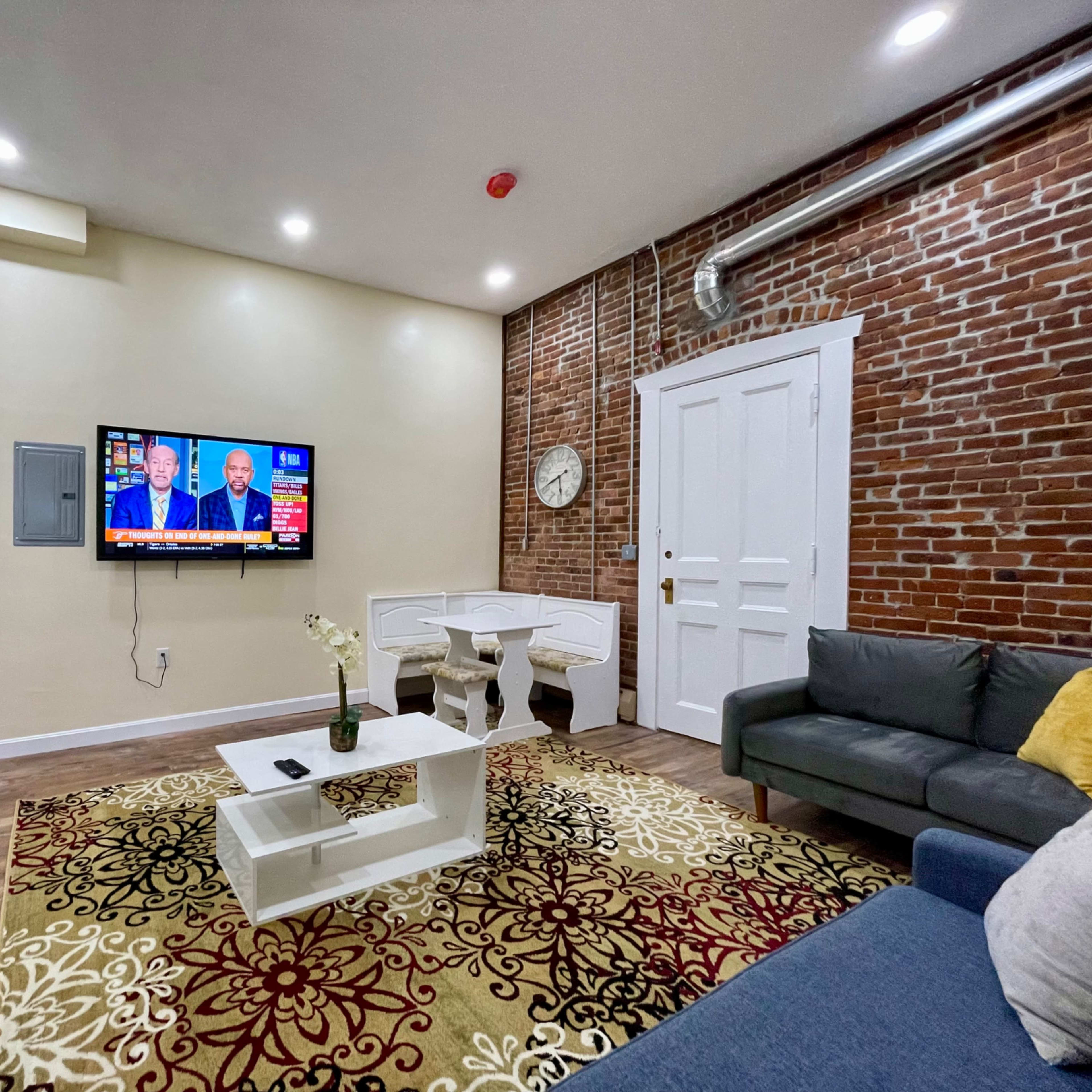 The living area features a television mounted on a wall with a brick backdrop, a gray sofa, a white coffee table, and a patterned area rug.