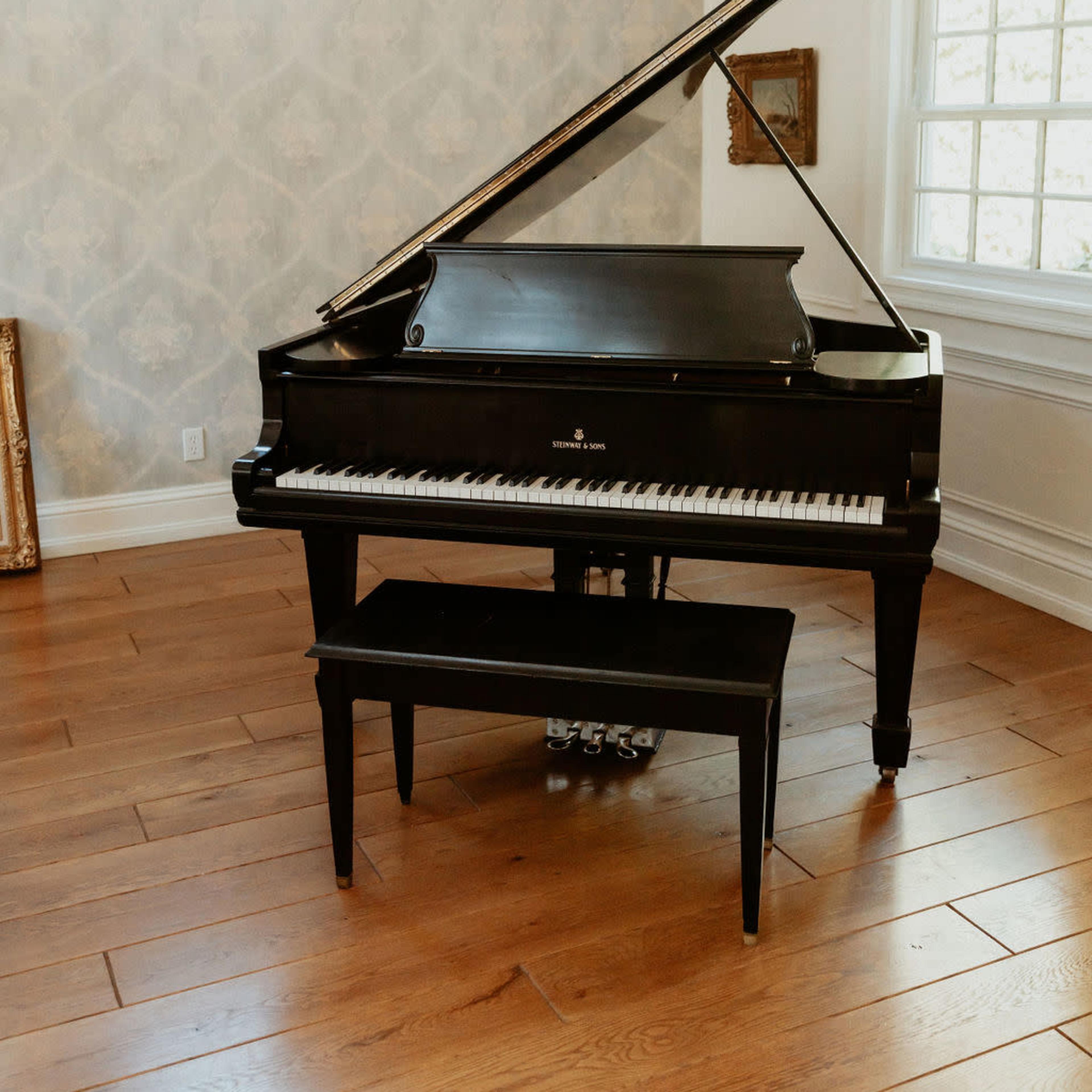 A black grand piano with an open lid and a matching bench is positioned on wooden flooring in a well-lit room with patterned wallpaper.