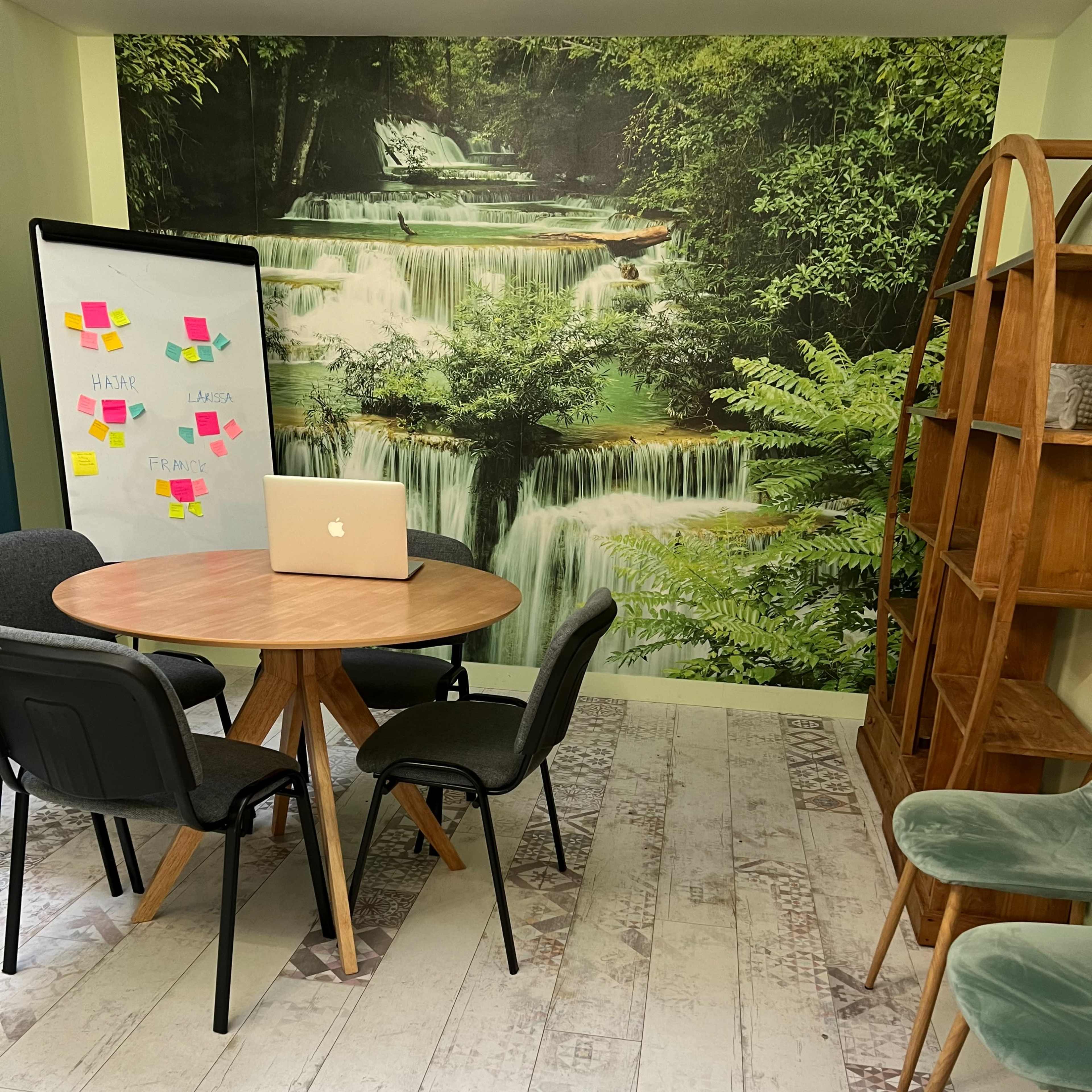 A small meeting area featuring a round table with chairs, a laptop on the table, a wall mural of a waterfall, and a wooden bookshelf against the wall.