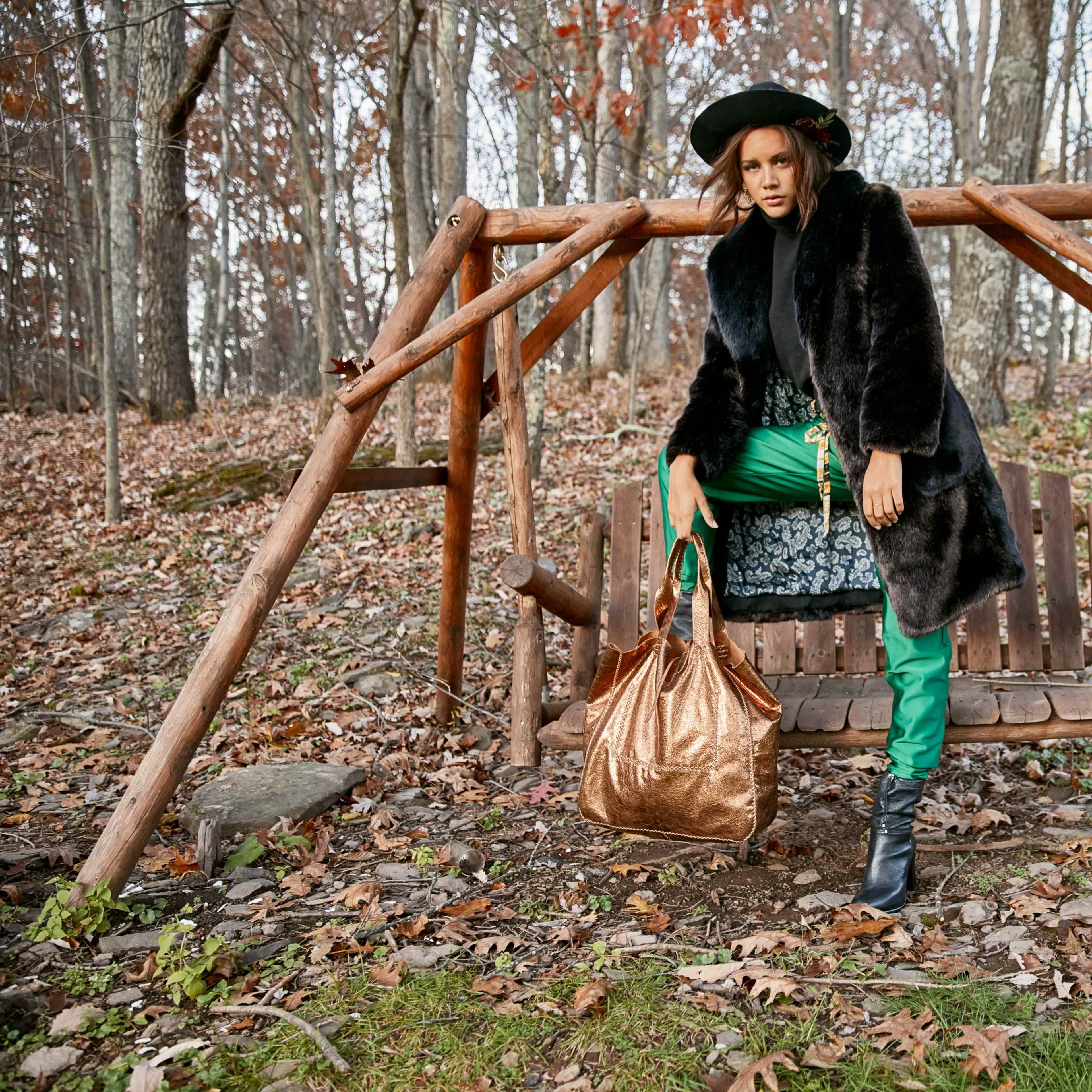 A person stands next to a wooden swing in a forested area, wearing a black fur coat and holding a large brown bag.