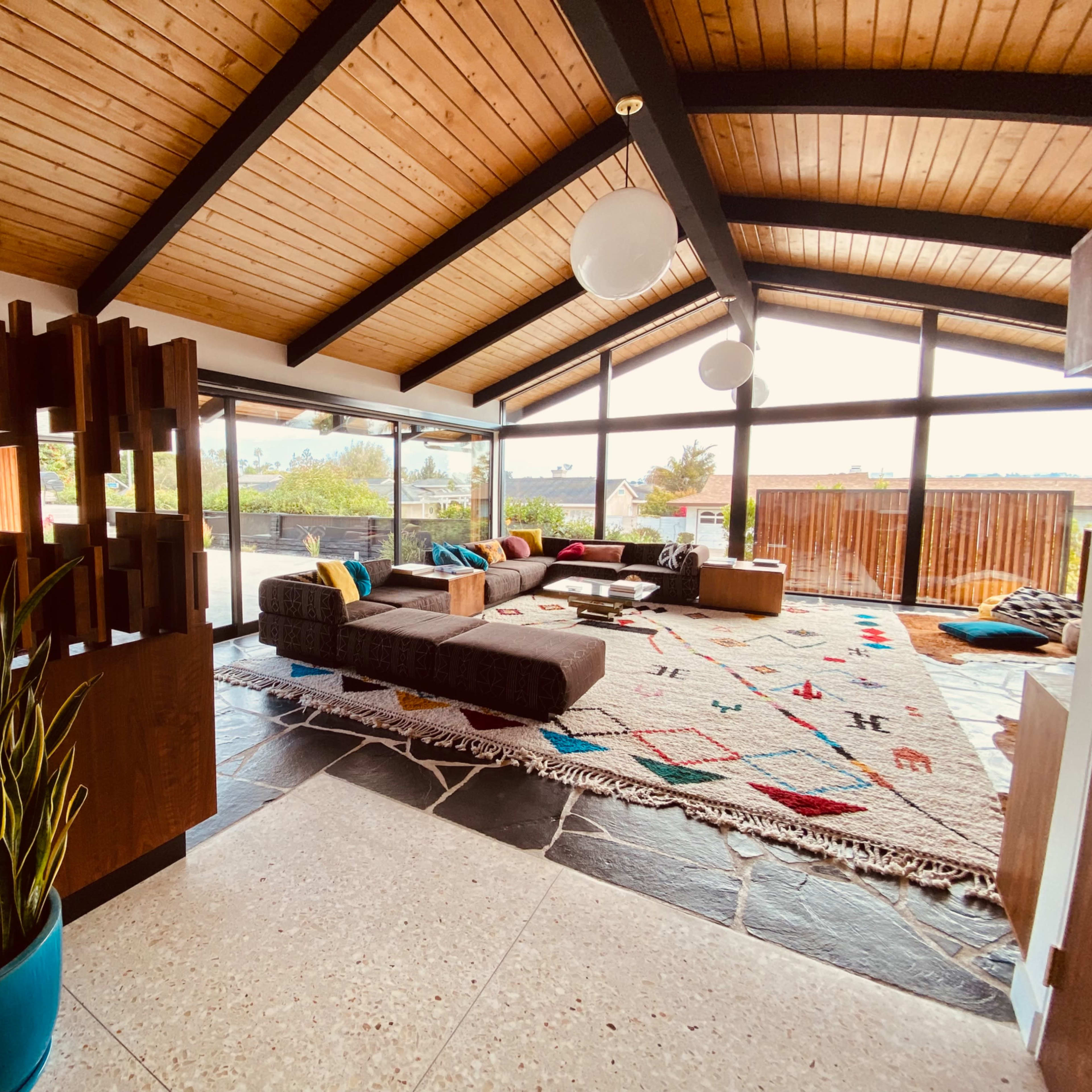 A modern living room with a high wooden ceiling, large windows, and a comfortable seating area arranged around a patterned rug.