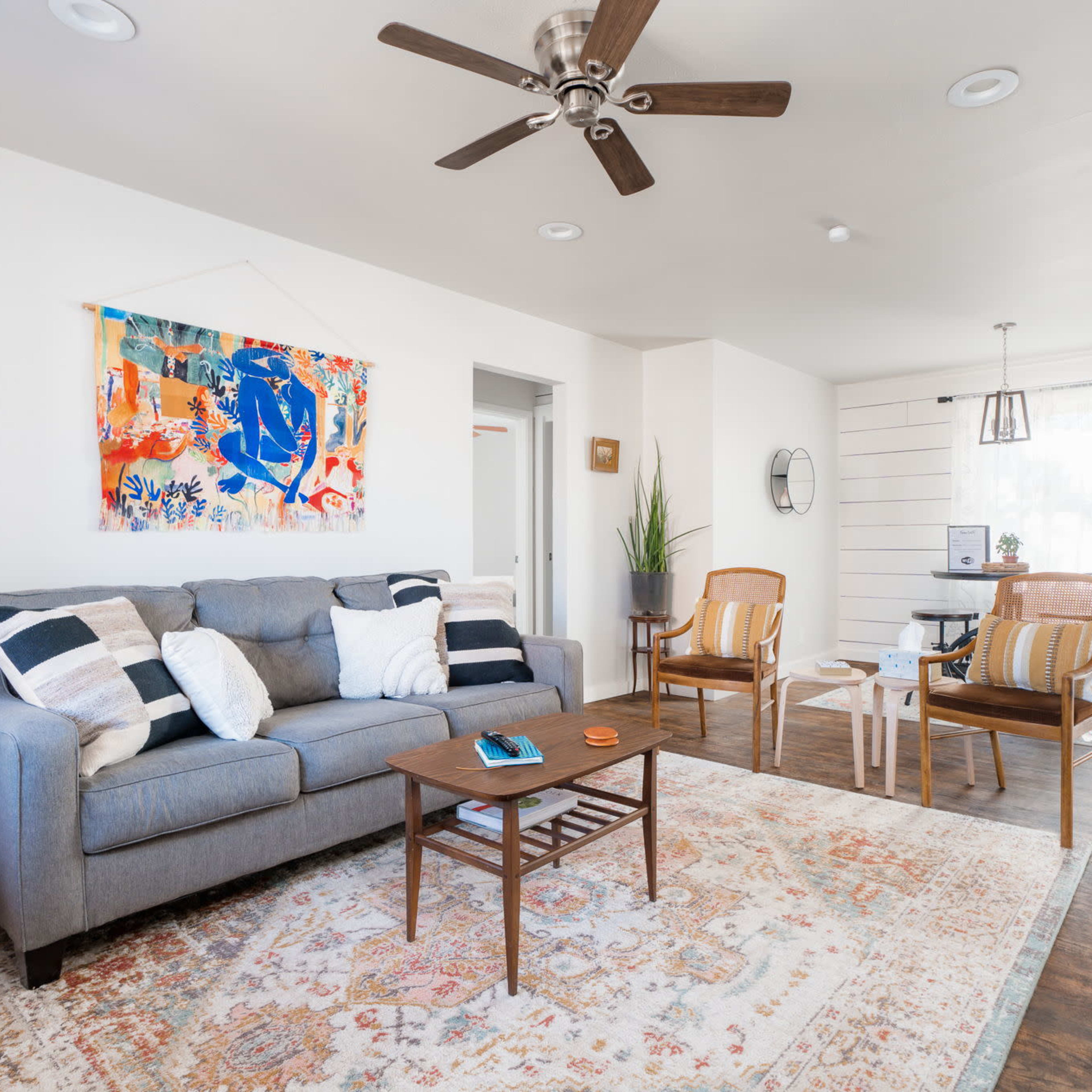A living room features a gray sofa, two wooden chairs, a small coffee table, and a patterned rug, with artwork hanging on the wall and a plant in the corner.