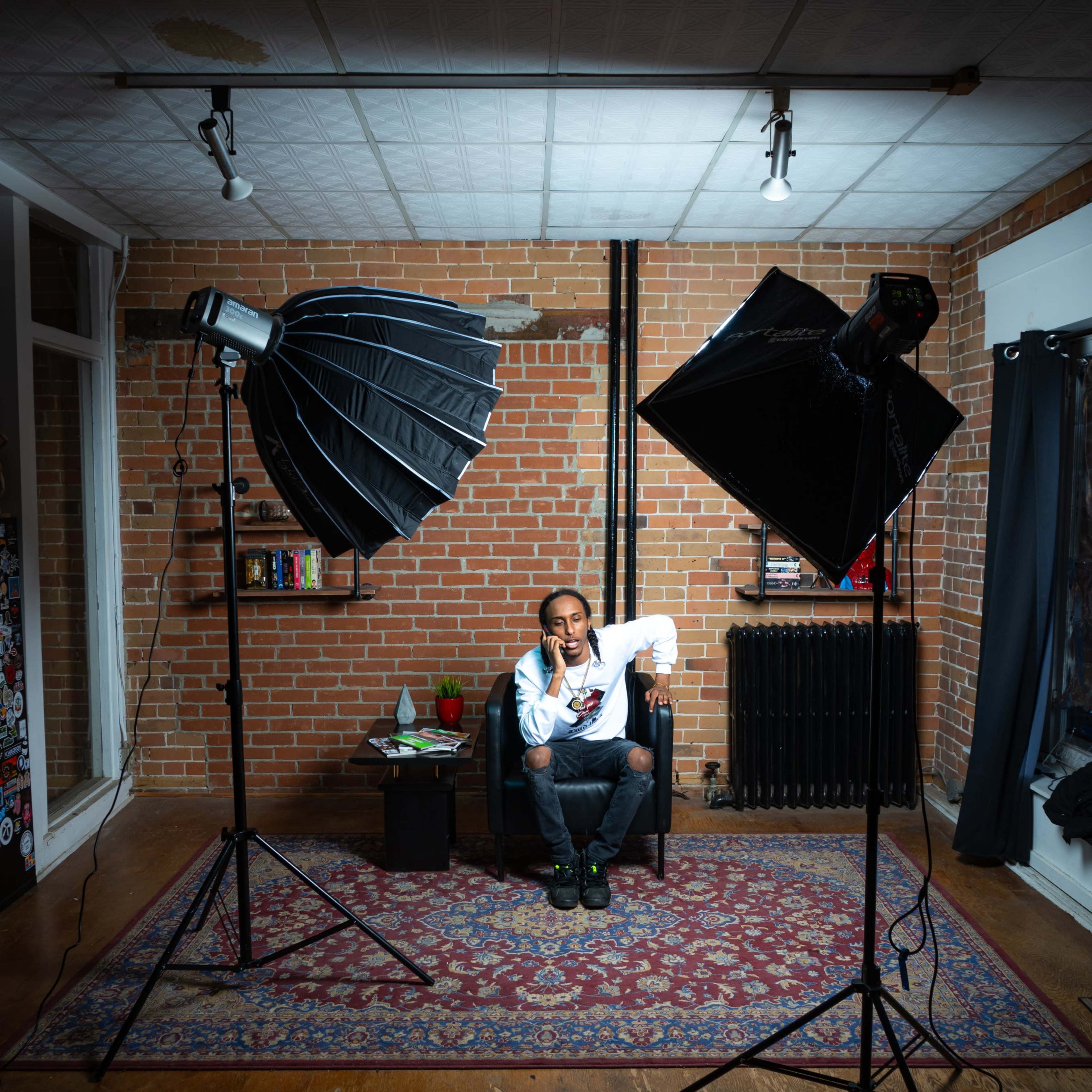 A person sits on a chair in a room with brick walls, surrounded by two softbox lights and a small table with a plant.