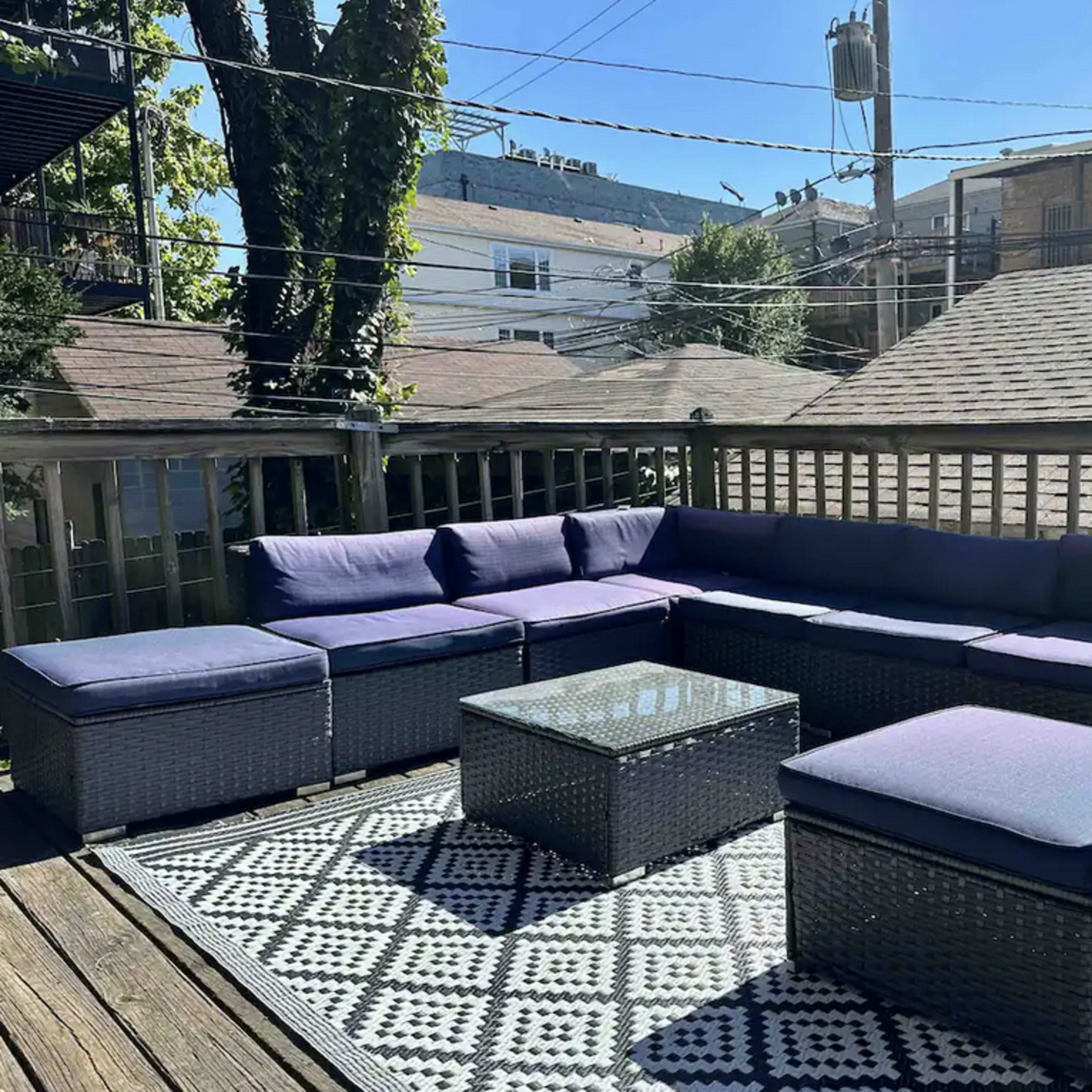 A patio deck featuring a large sectional sofa, a glass-top table, and a patterned rug in an outdoor setting with nearby houses in the background.
