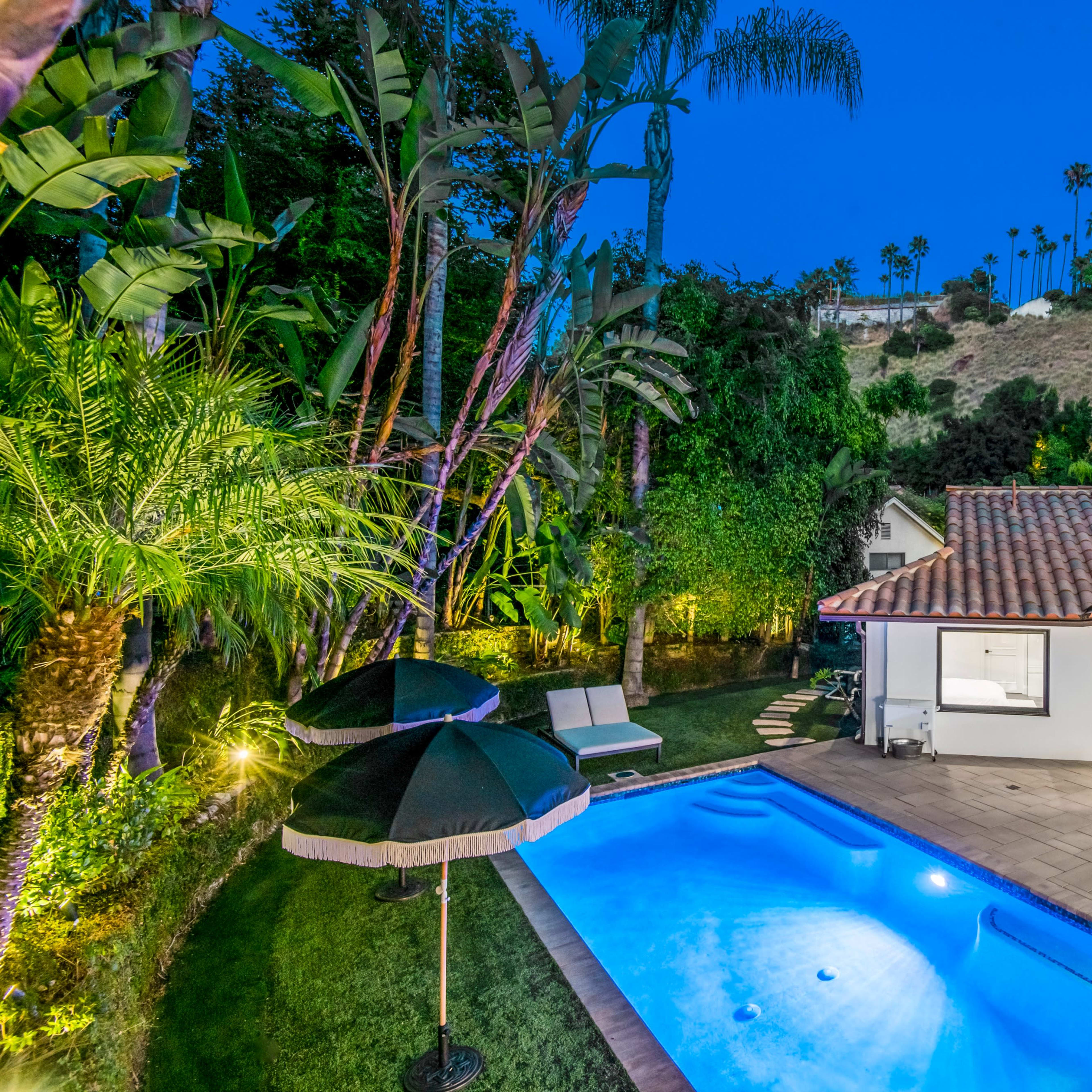 A landscaped backyard with a swimming pool, patio furniture, and tropical plants surrounded by palm trees at dusk.