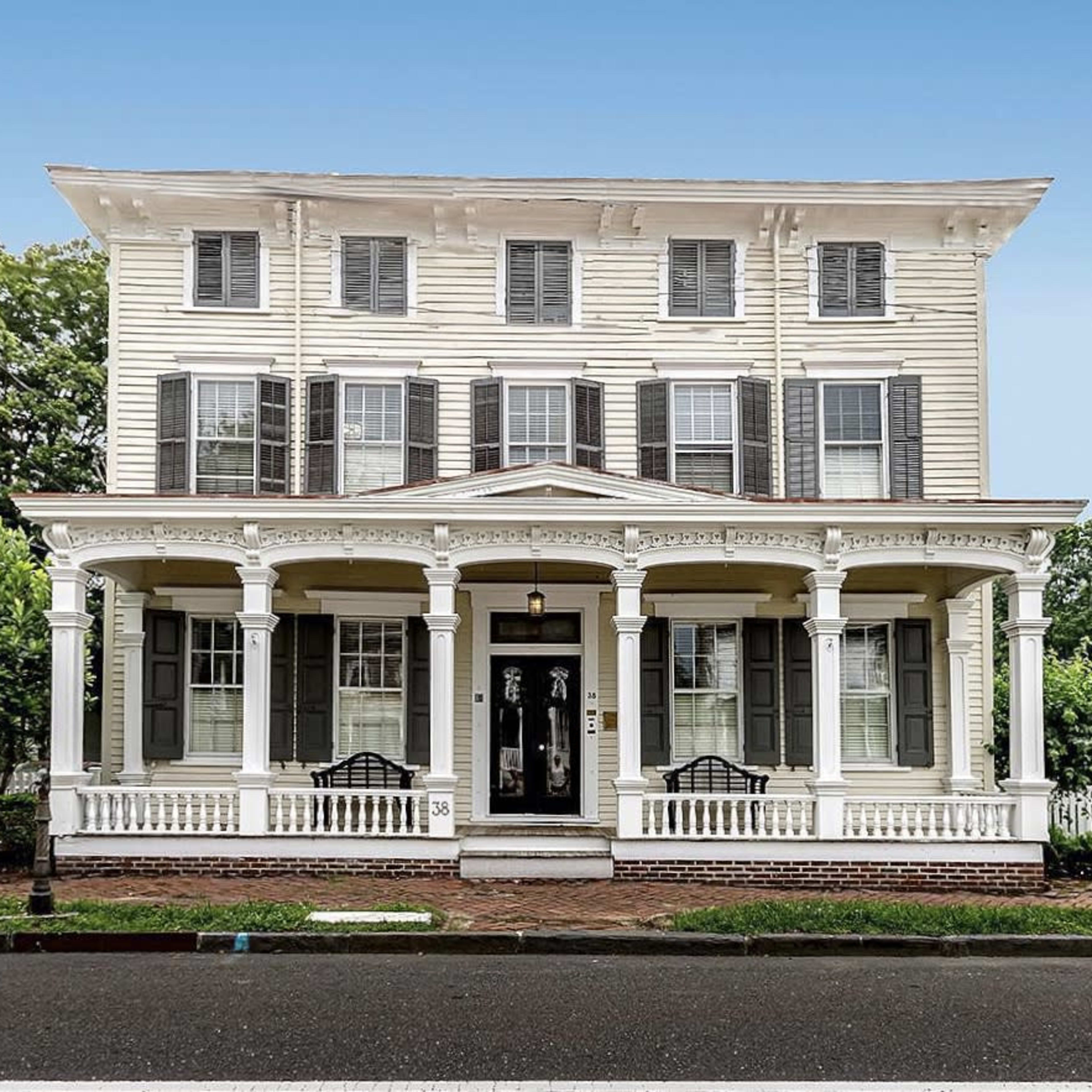 A three-story white house with a front porch supported by columns, featuring multiple windows with shutters.