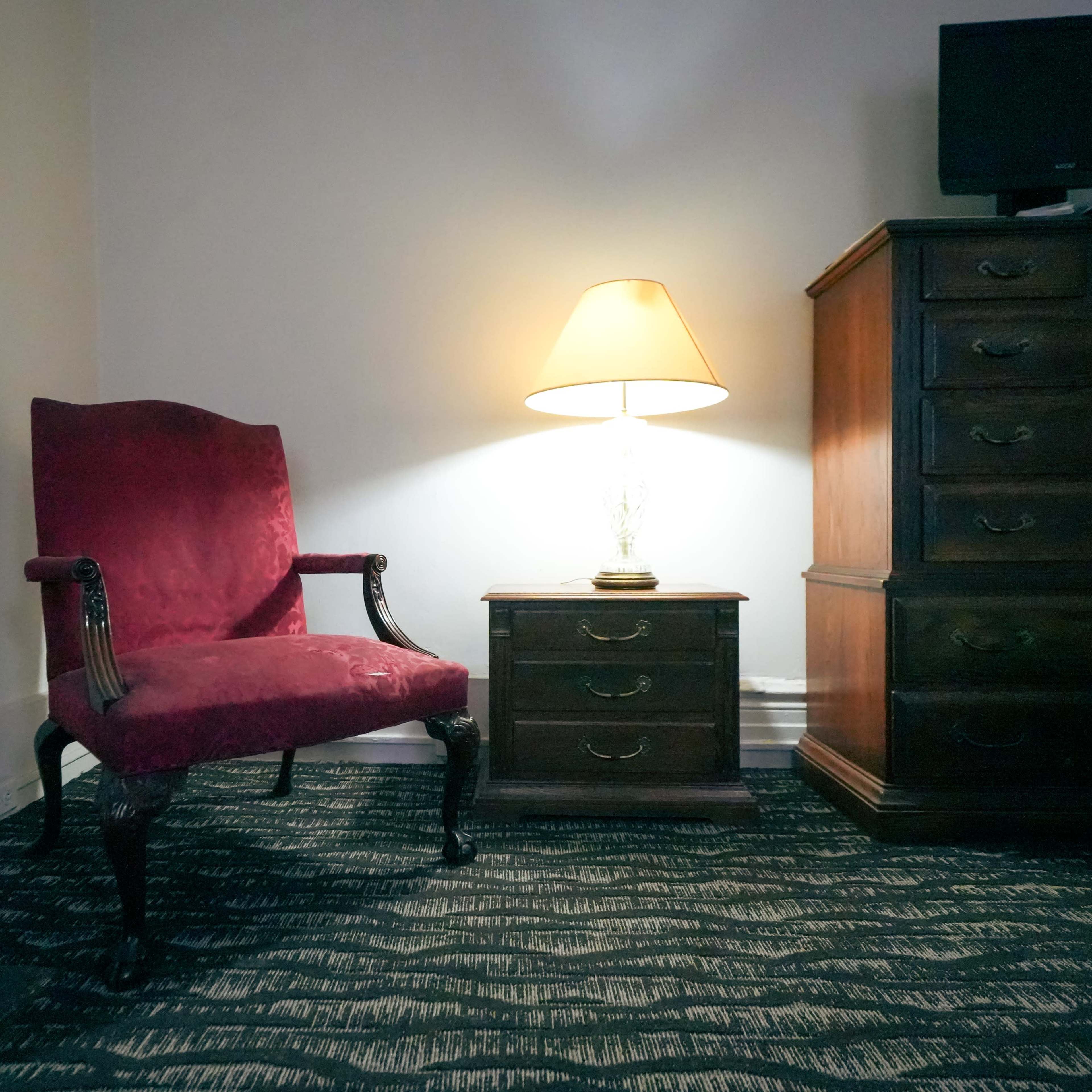 A red upholstered armchair sits next to a nightstand with a lamp, while a wooden dresser is positioned against the wall in a softly lit room.