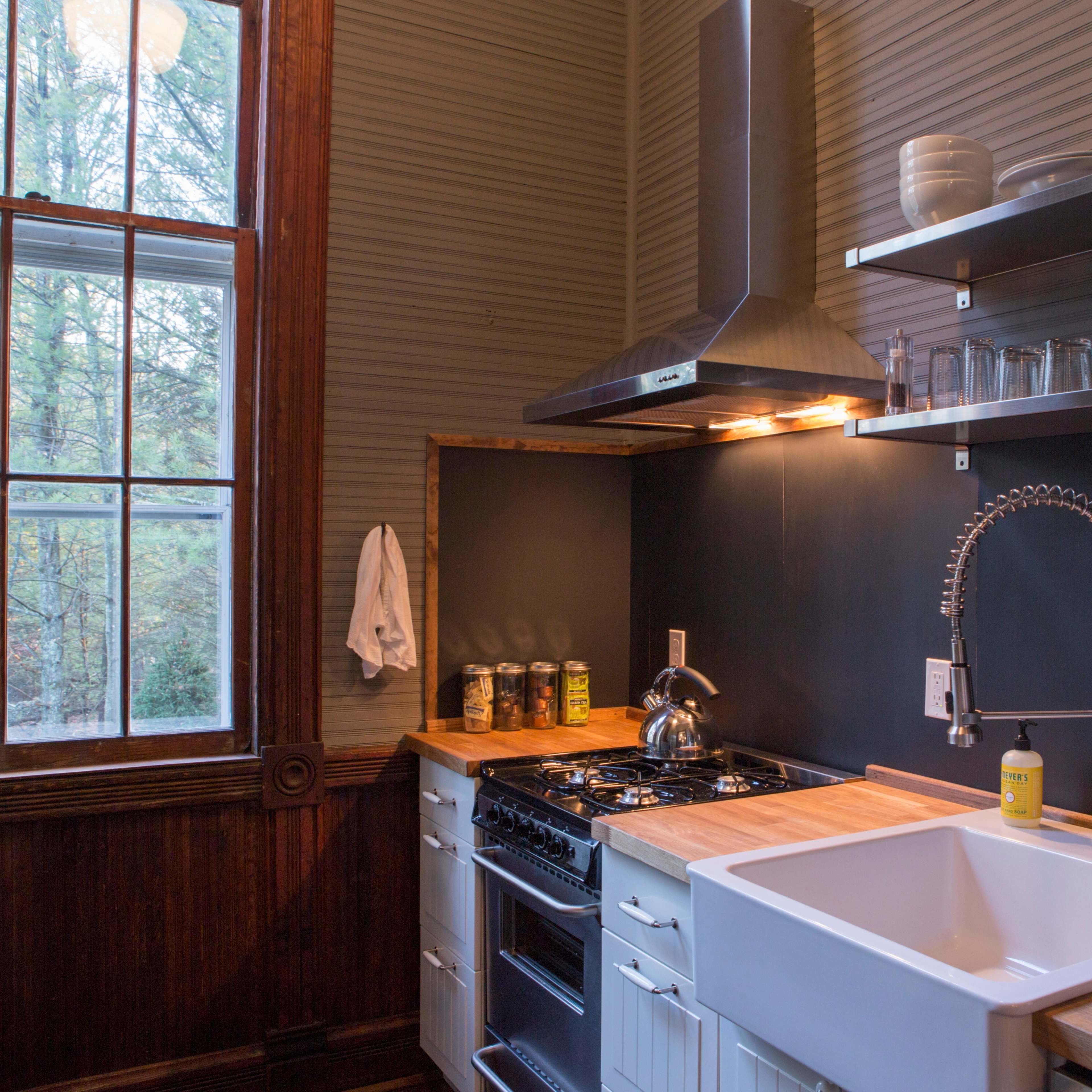 A contemporary kitchen features a large sink, a stovetop with a hood, and shelves displaying glassware and jars, with a large window providing natural light.
