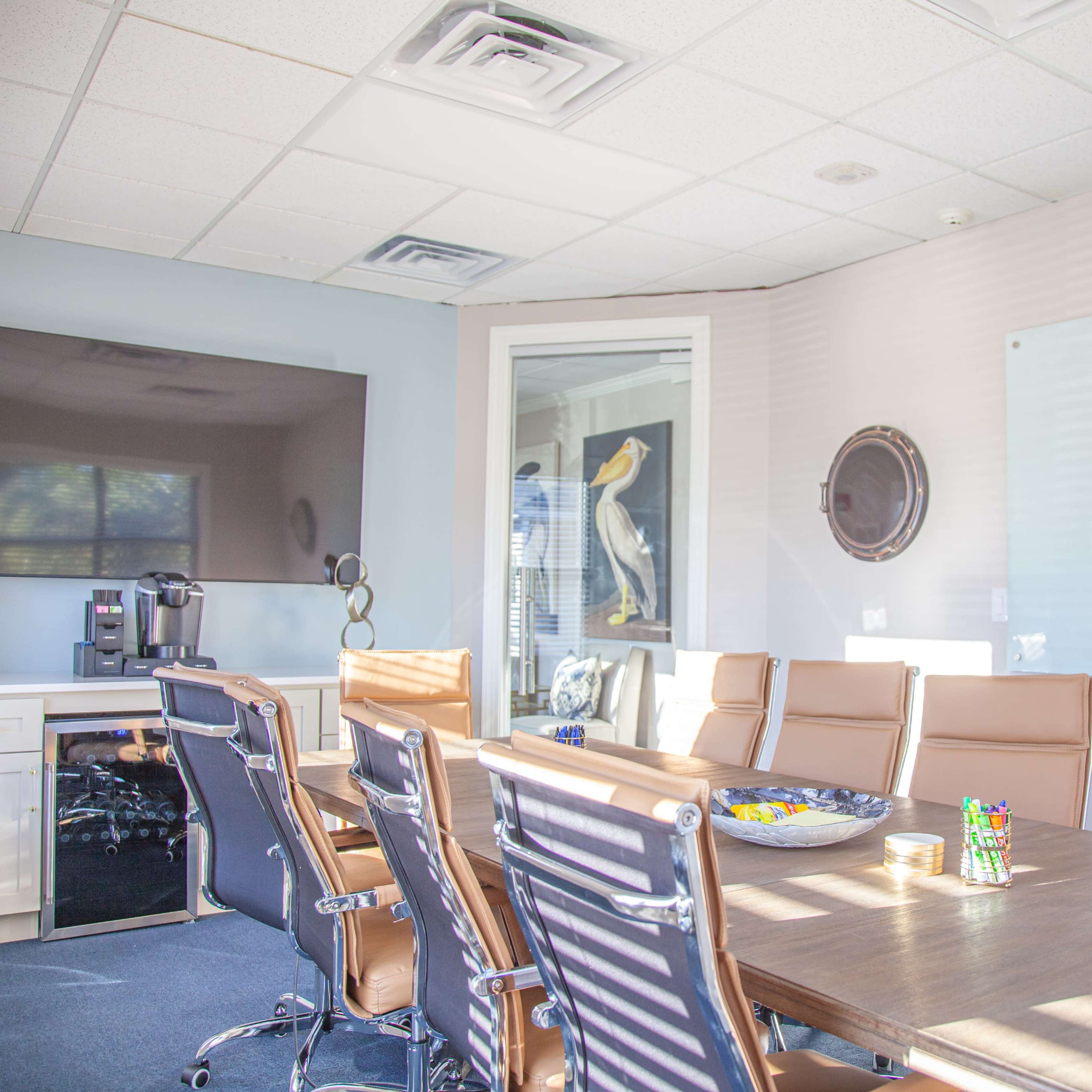 A modern conference room features a long wooden table surrounded by leather chairs, a large television screen on the wall, and a kitchenette area with a coffee machine.