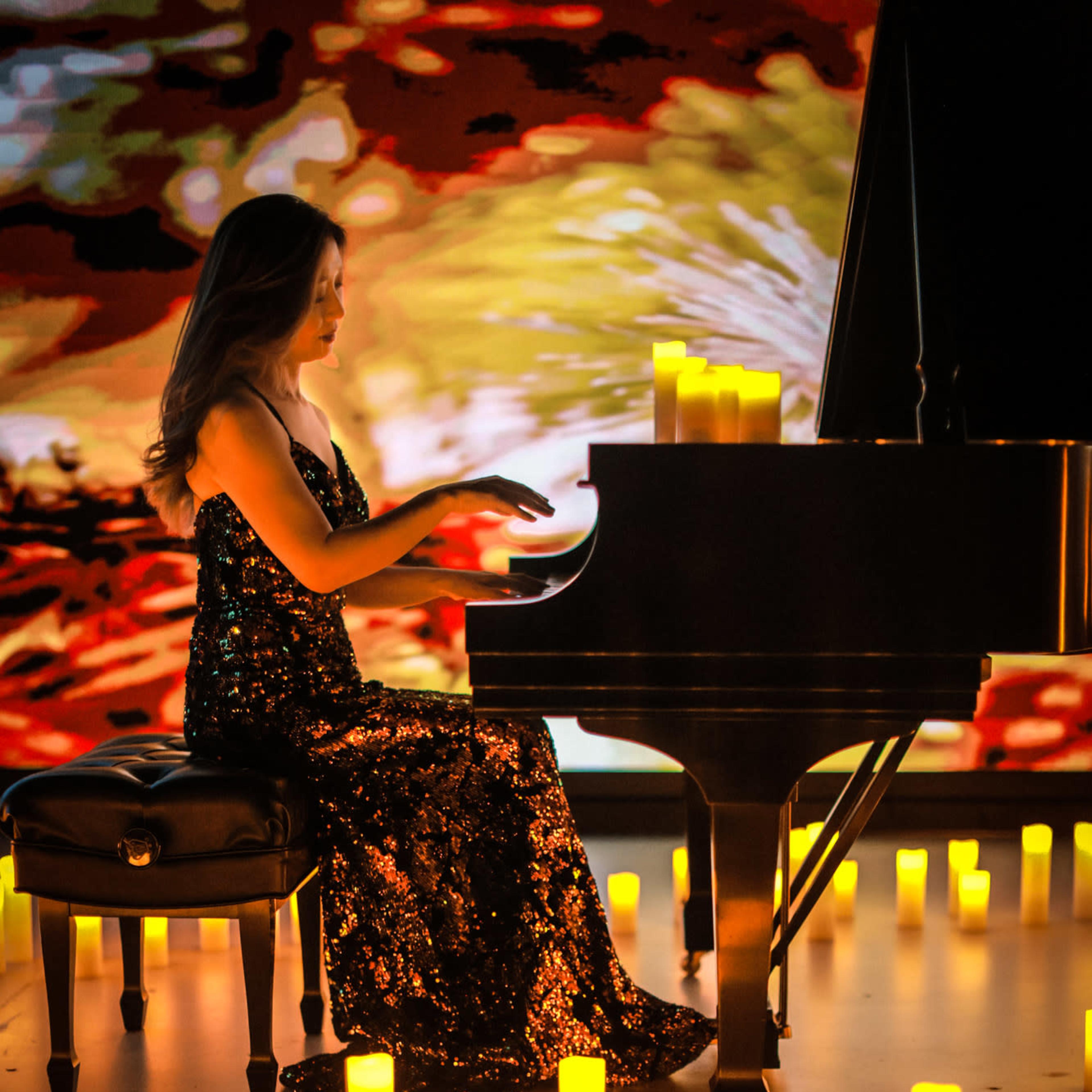A woman in a sequined dress plays a piano surrounded by glowing candles on the floor.