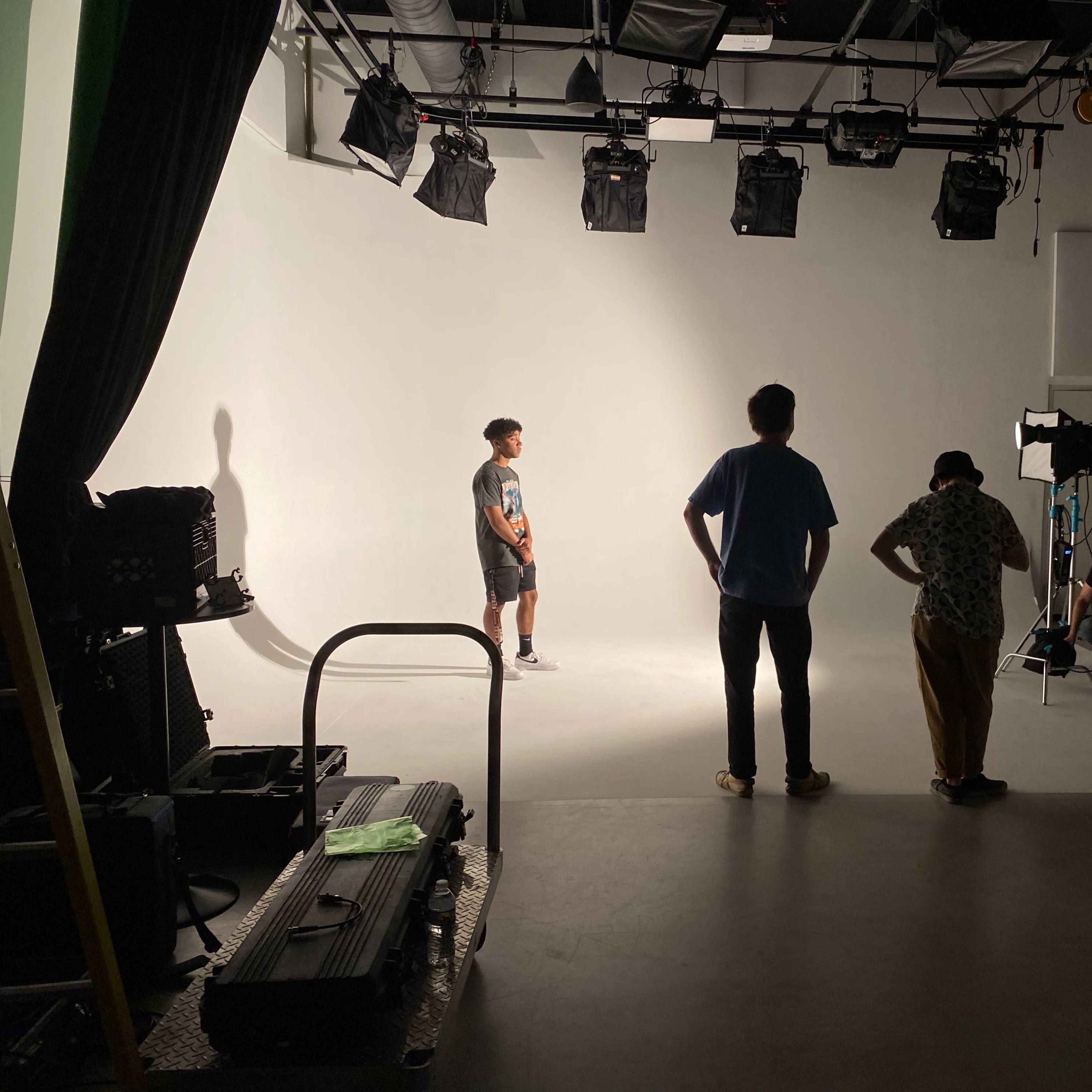 A model stands on a well-lit stage while a small crew observes from the side, with cameras and equipment set up for a shoot.