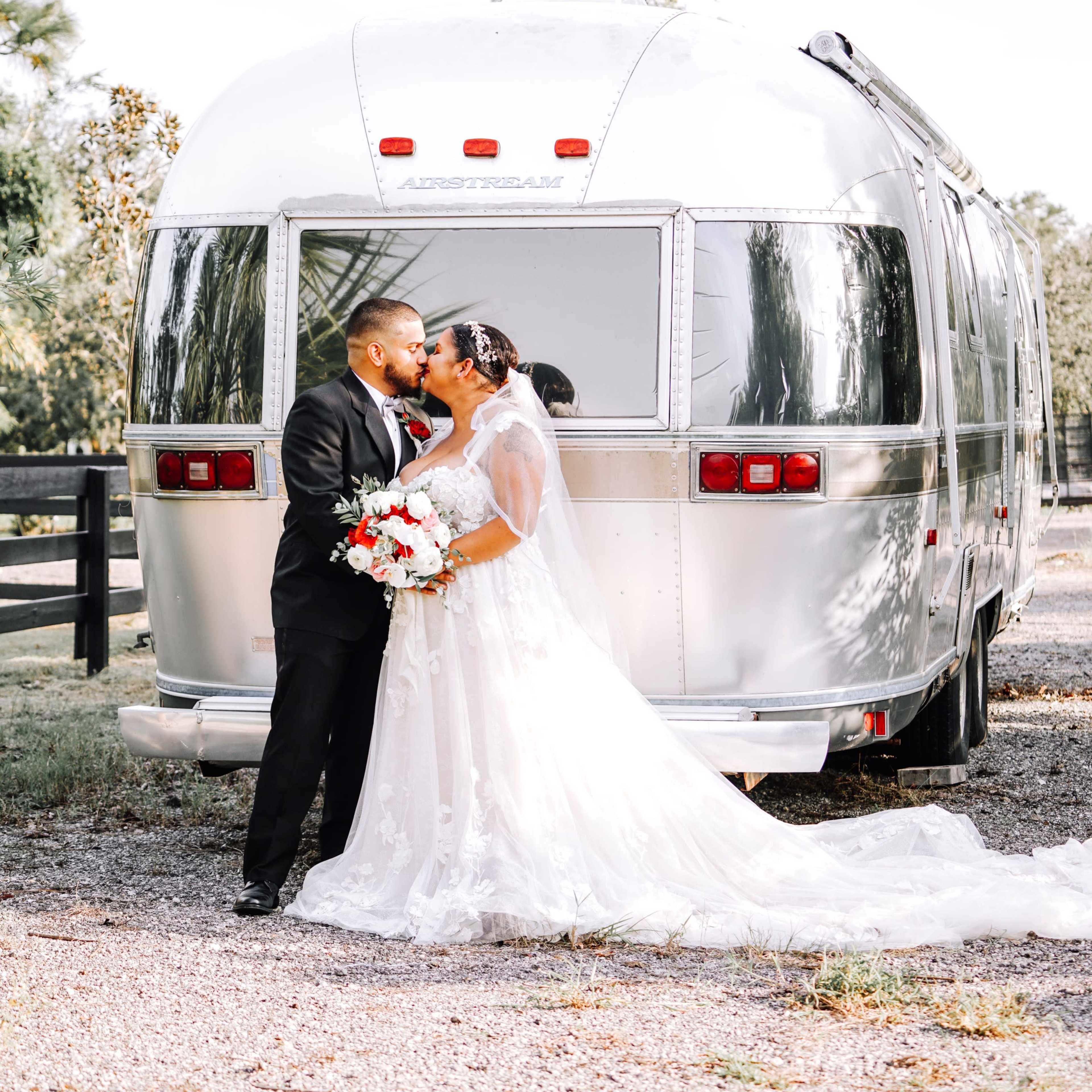 A couple dressed in formal wedding attire shares a kiss in front of a vintage Airstream trailer.