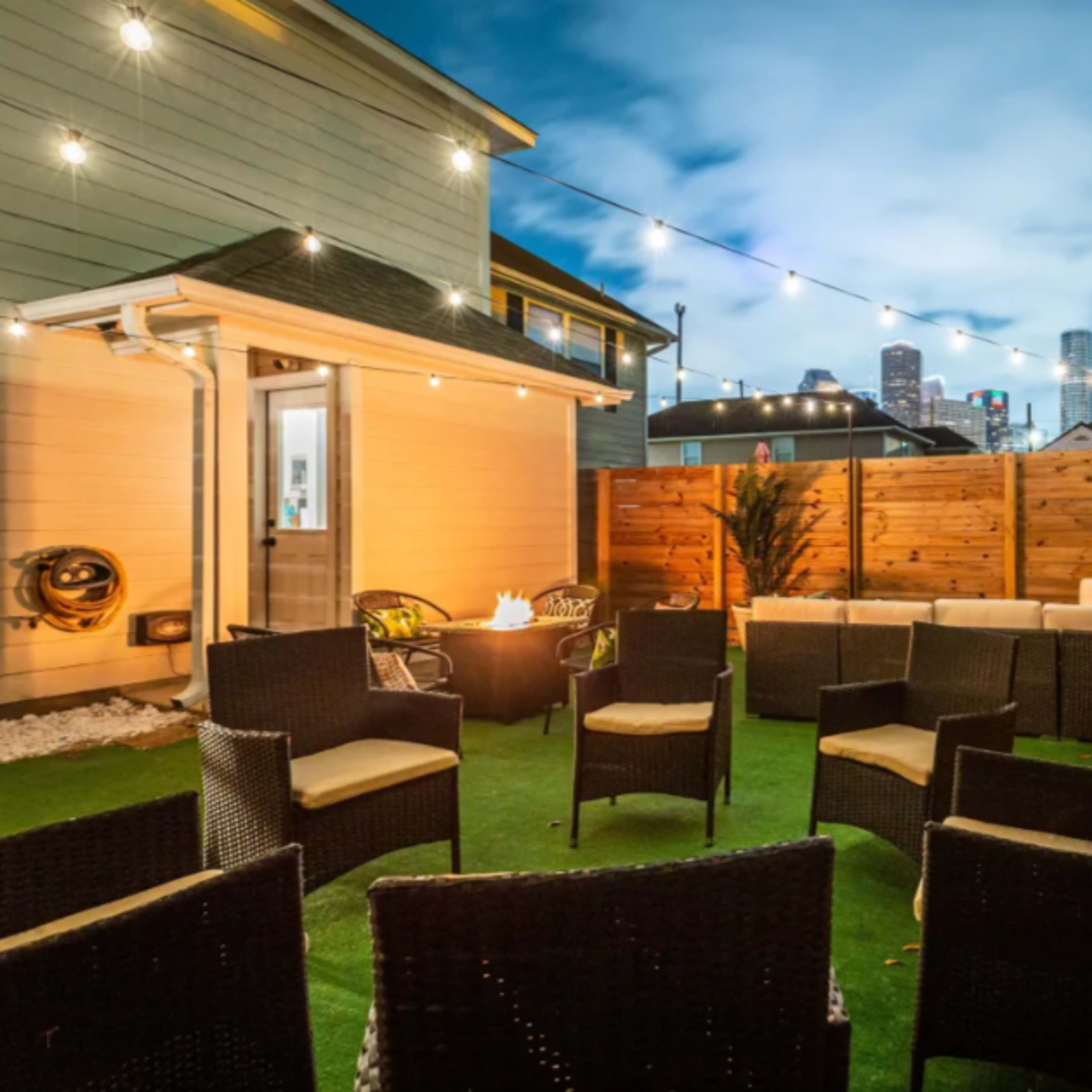 A cozy outdoor seating area with wicker chairs arranged around a central fire pit, set against a backdrop of a city skyline at dusk.