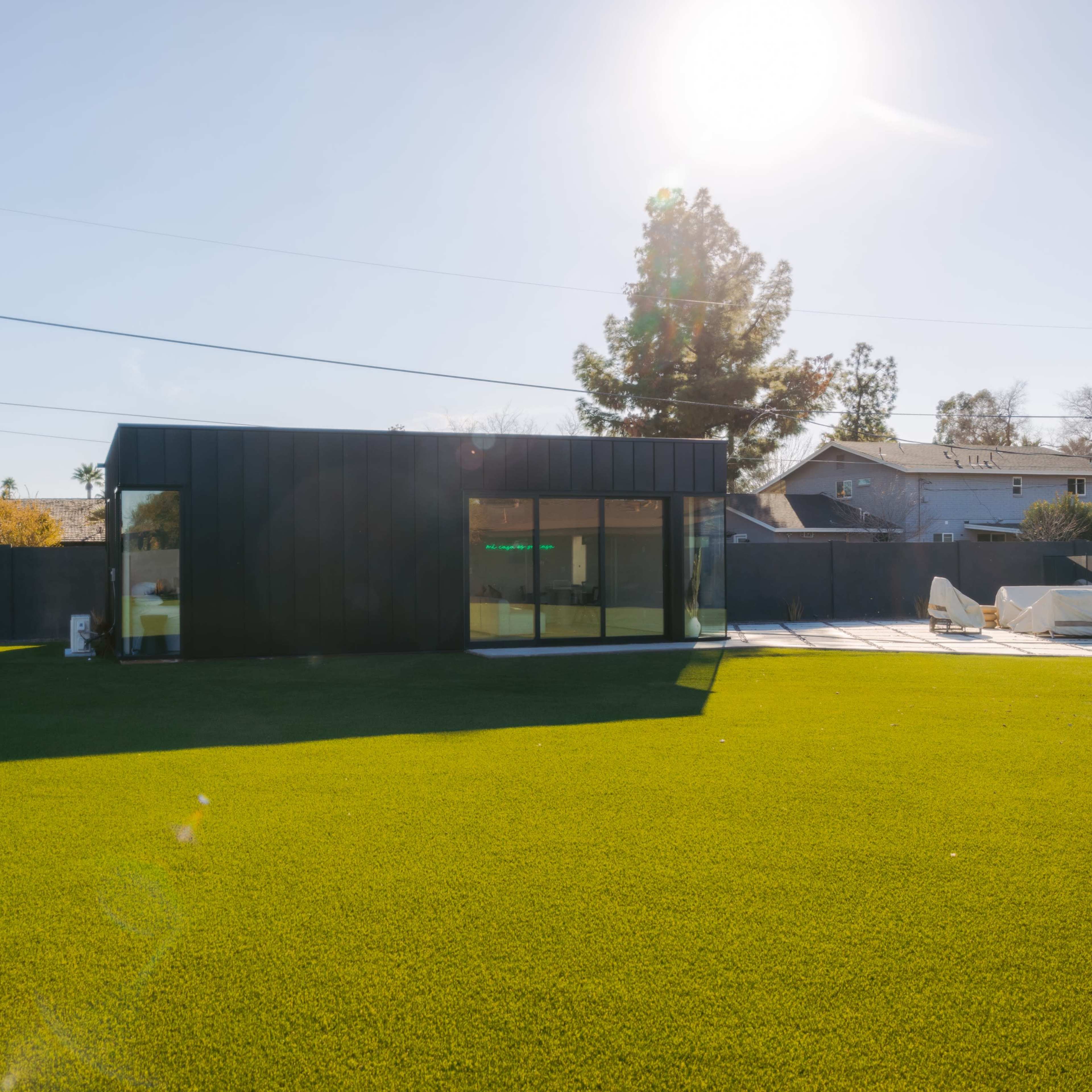 A modern black structure with large windows is surrounded by a green lawn under a clear sky.