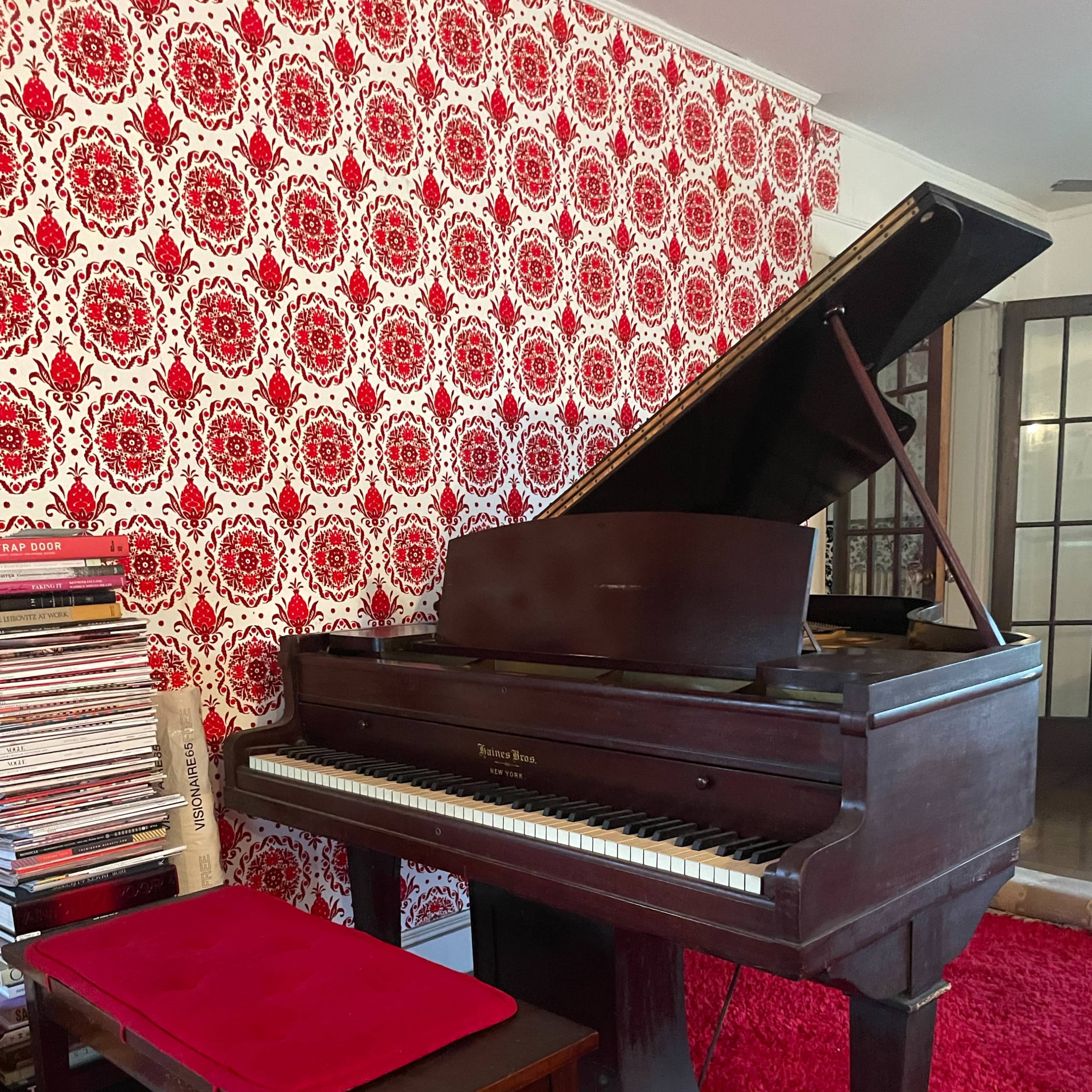 A grand piano with an open lid is positioned beside a stack of records against a wall covered in red patterned wallpaper.