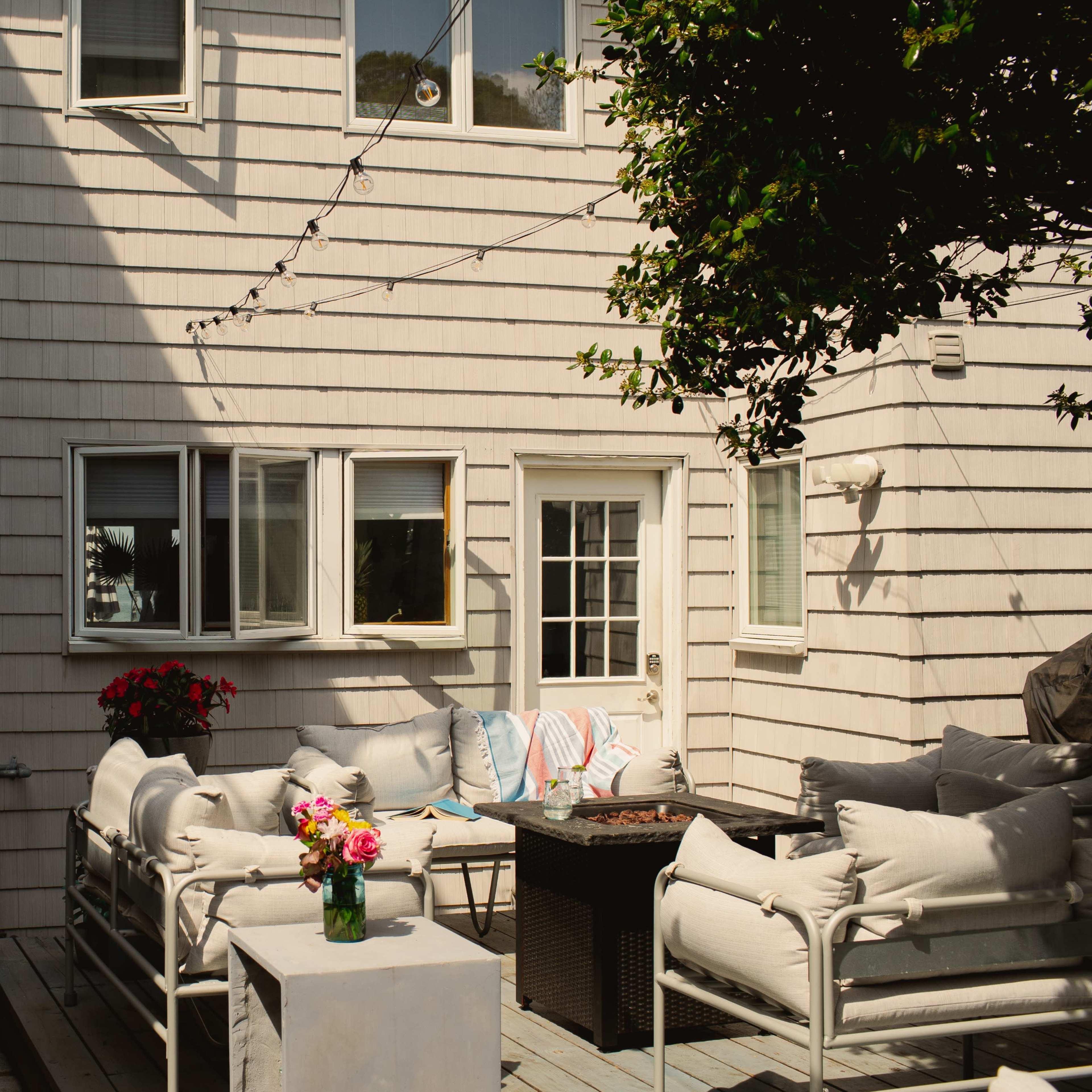 A well-furnished outdoor patio area next to a waterfront, with seating, a table, and potted flowers against the backdrop of a wooden house.