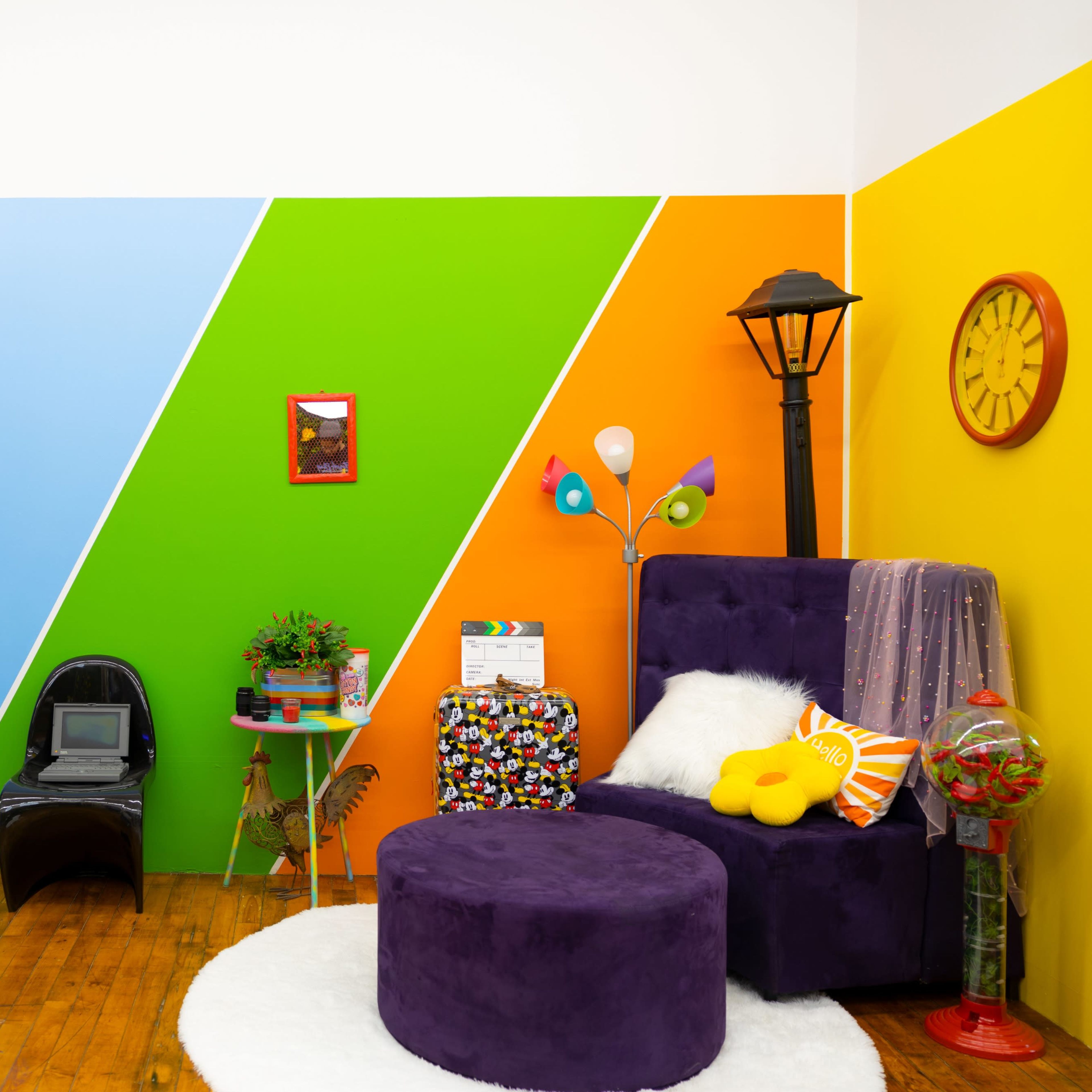 A vibrant room with colorful walls in pink, blue, green, and orange, featuring a purple chair, a round purple ottoman, a large mirror, decorative plants, and a lamp.