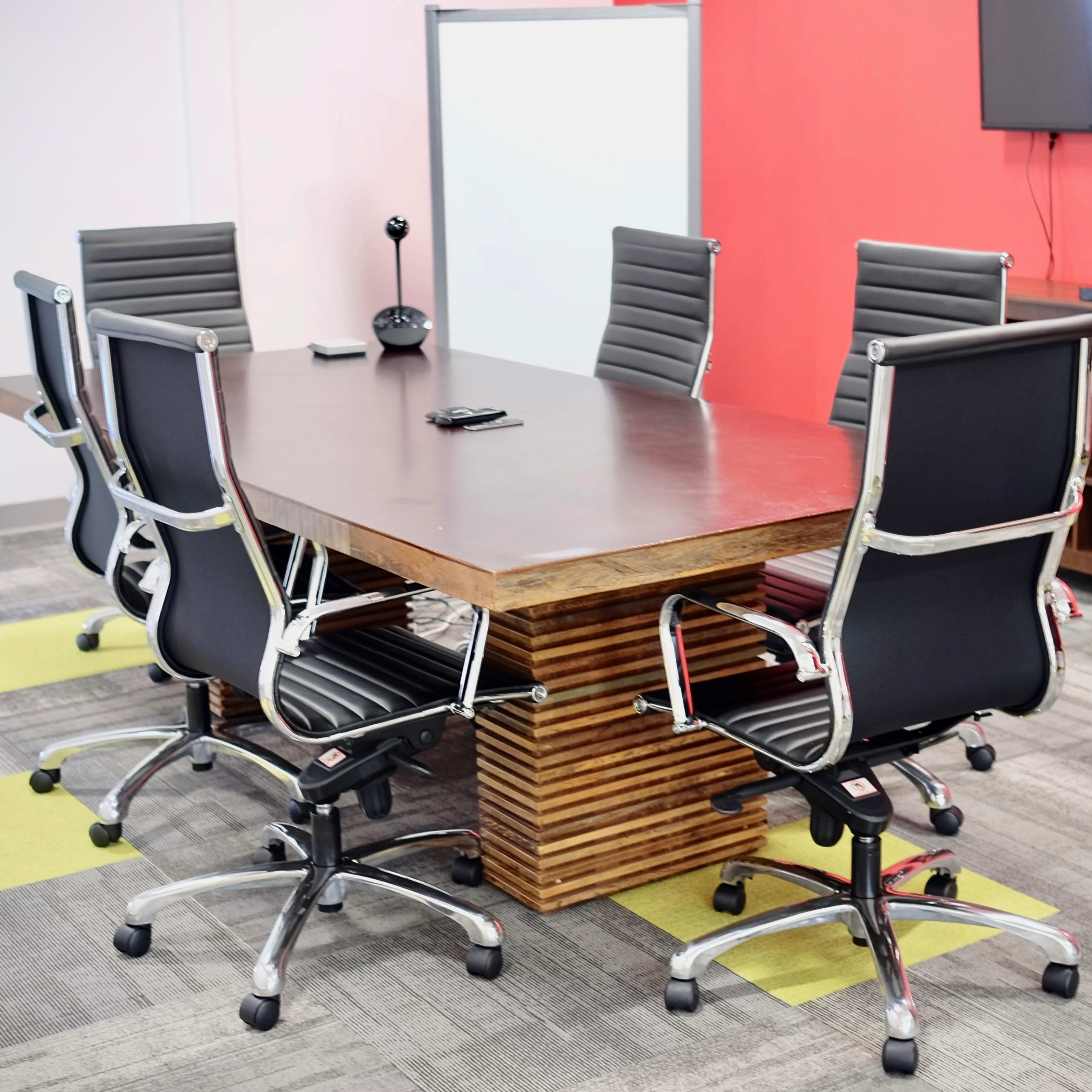 A modern conference room features a rectangular wooden table surrounded by six black office chairs, with a television mounted on the wall and a whiteboard in the corner.