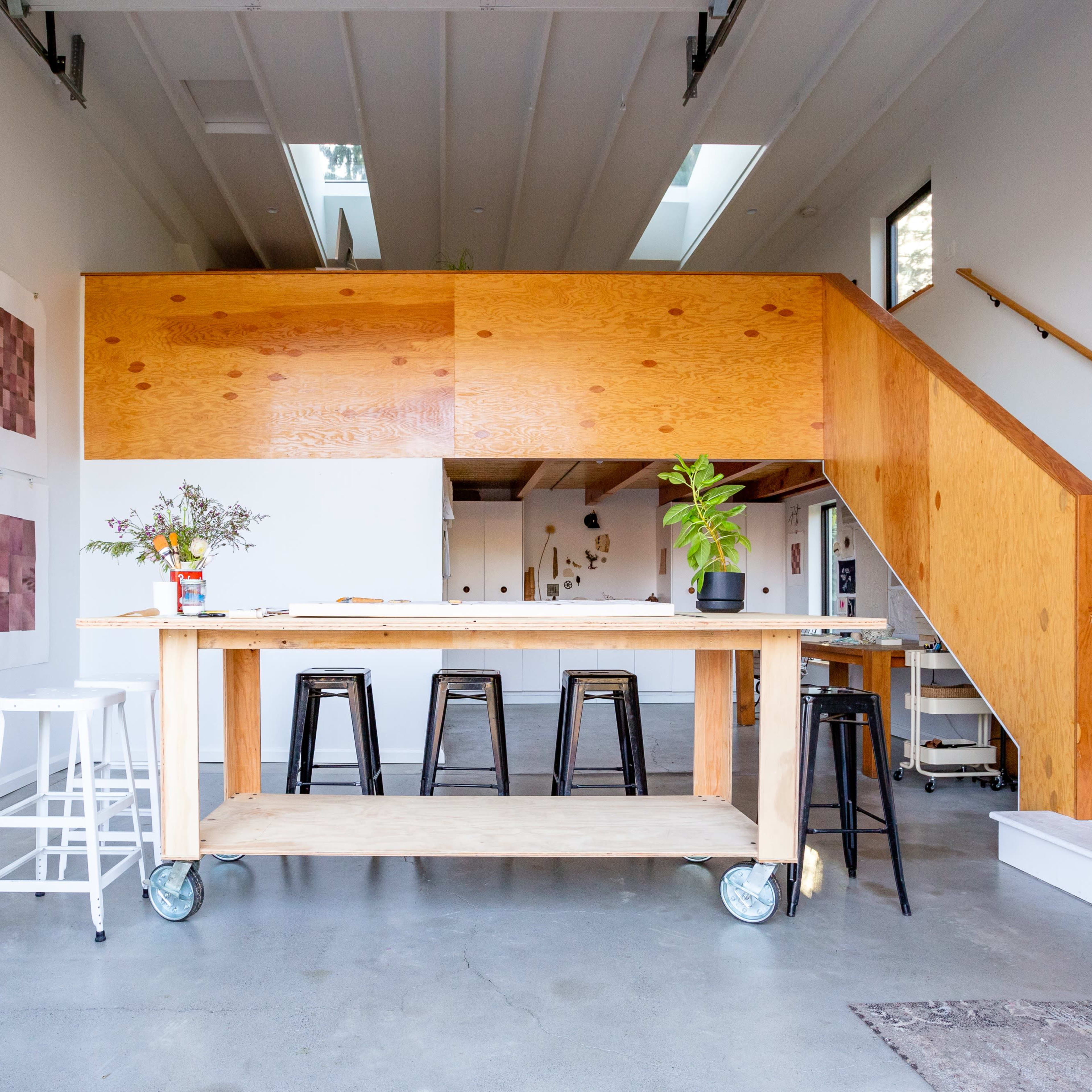 A modern kitchen area with a wooden island on wheels, black metal stools, and a staircase in the background.
