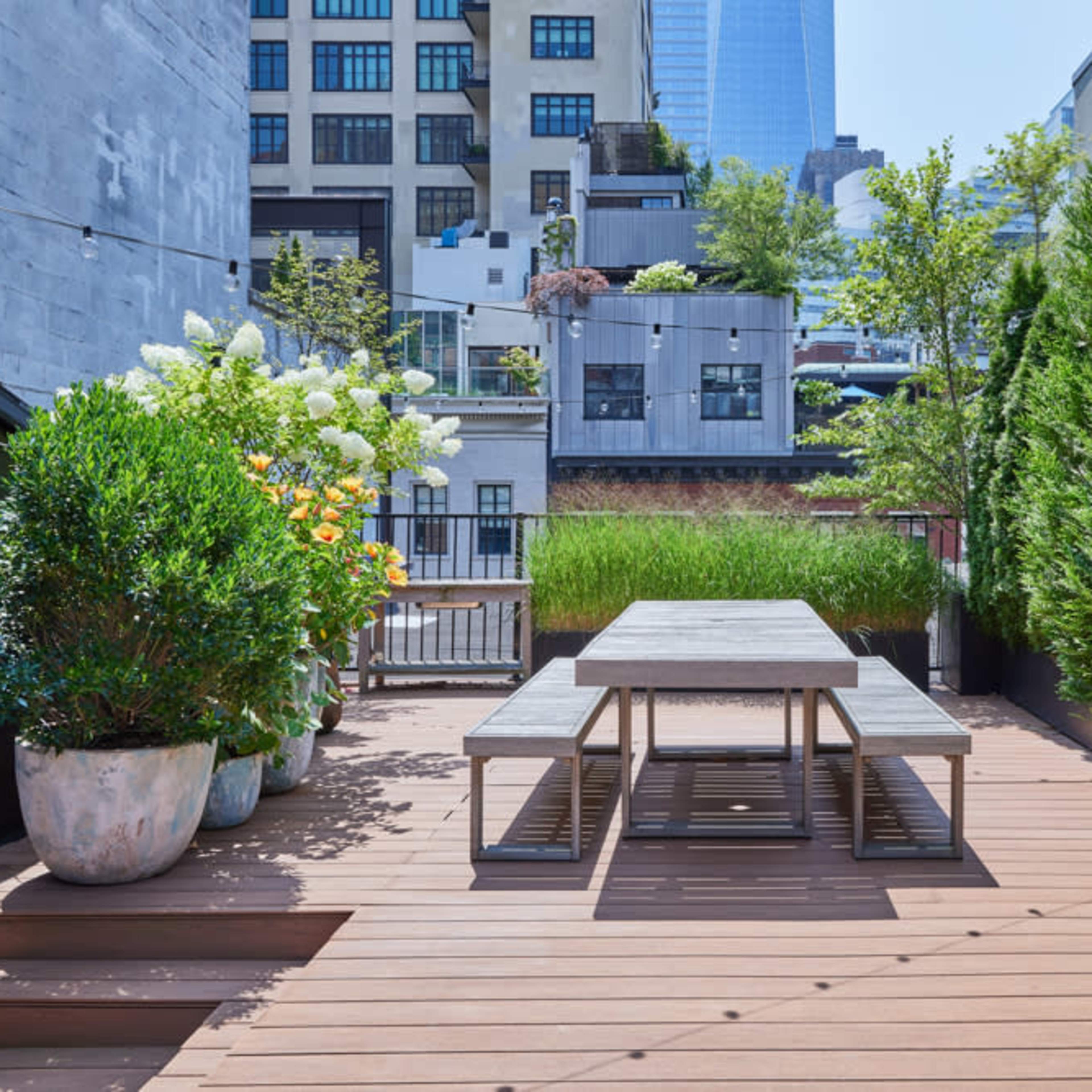 A rooftop terrace featuring wooden decking, a table with benches, and large planters filled with greenery.