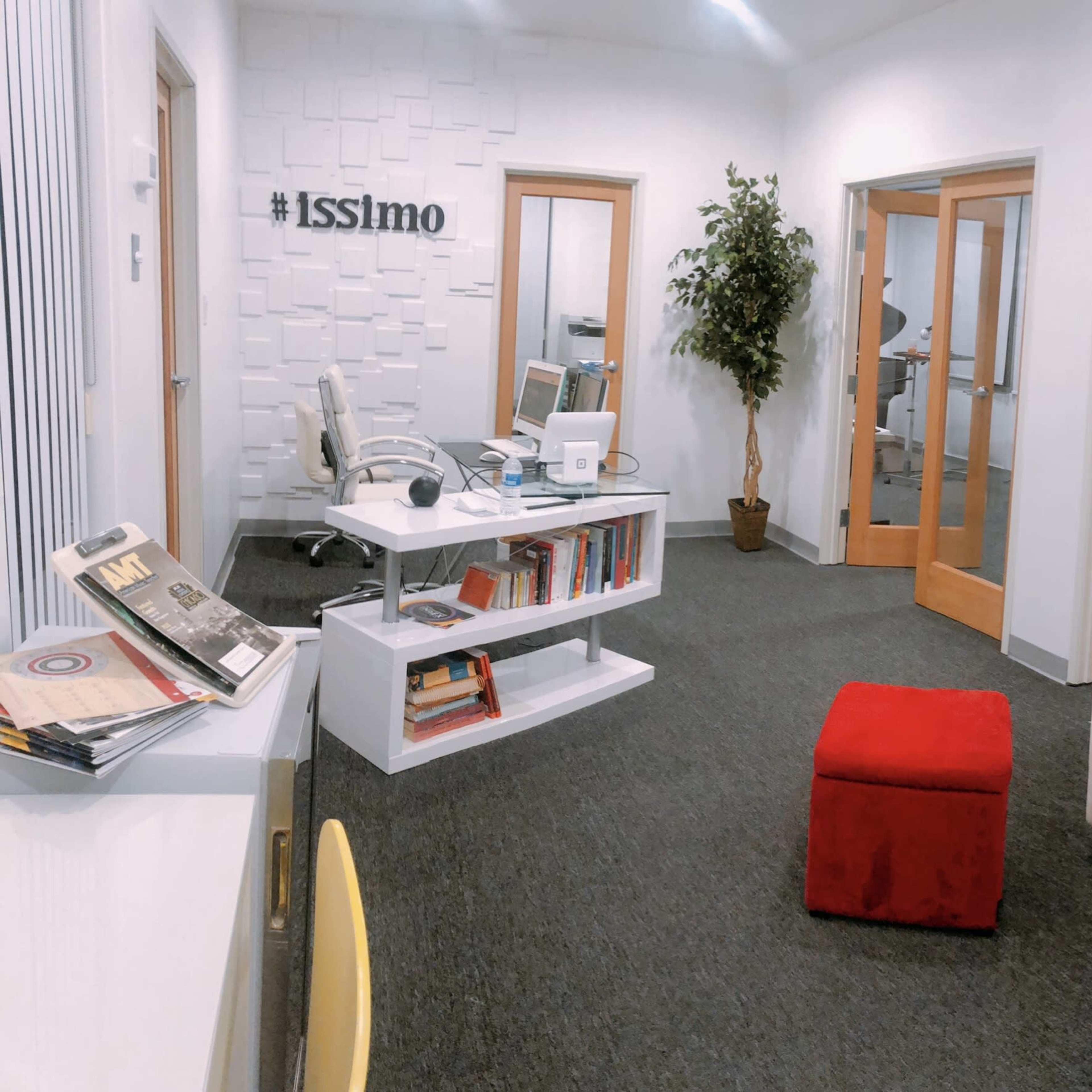 A modern office space featuring a white sofa, a small red ottoman, a desk with books, and a potted plant against a wall with the hashtag "#Isslimo."