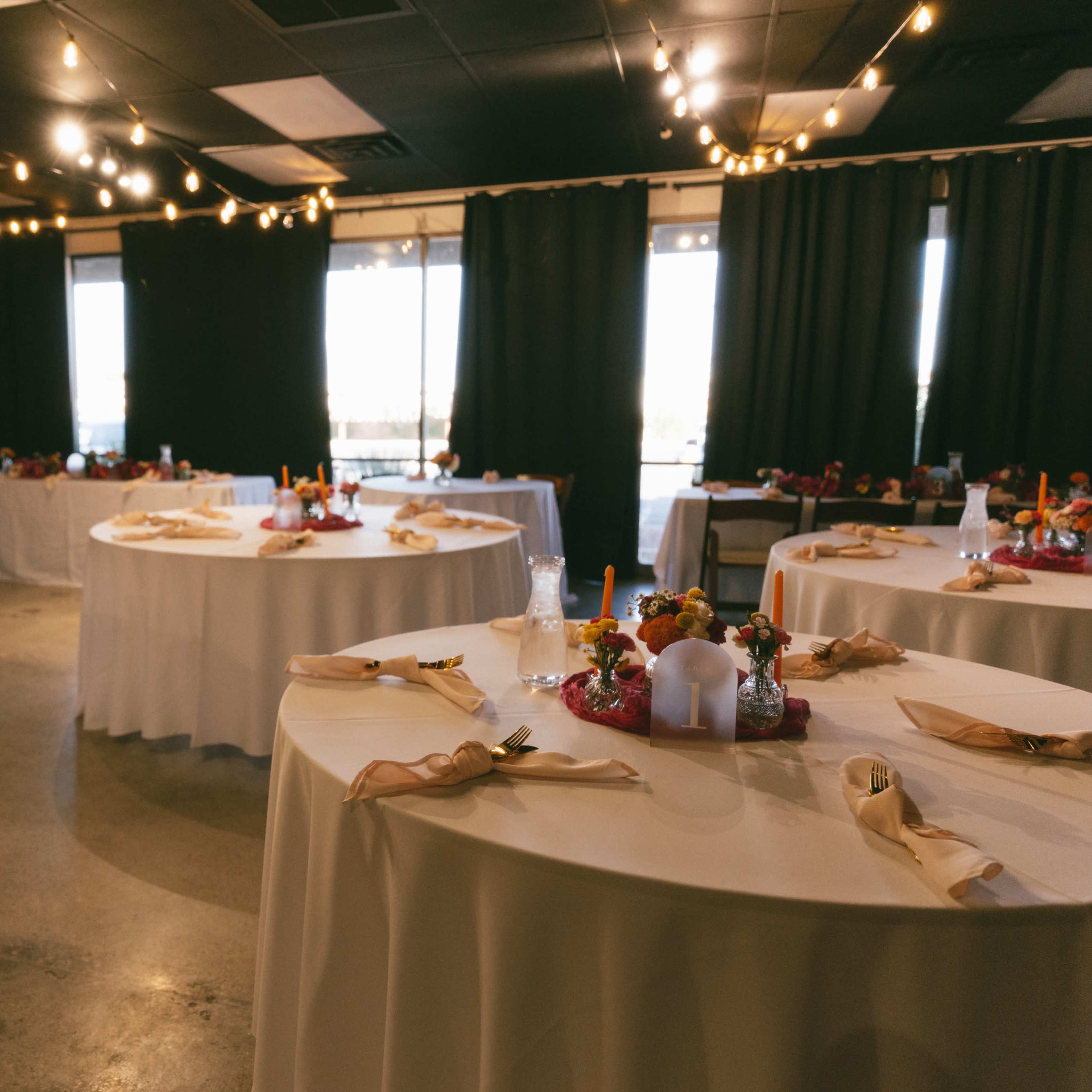 A banquet room with several circular tables set with white tablecloths and colorful centerpieces, illuminated by decorative string lights.