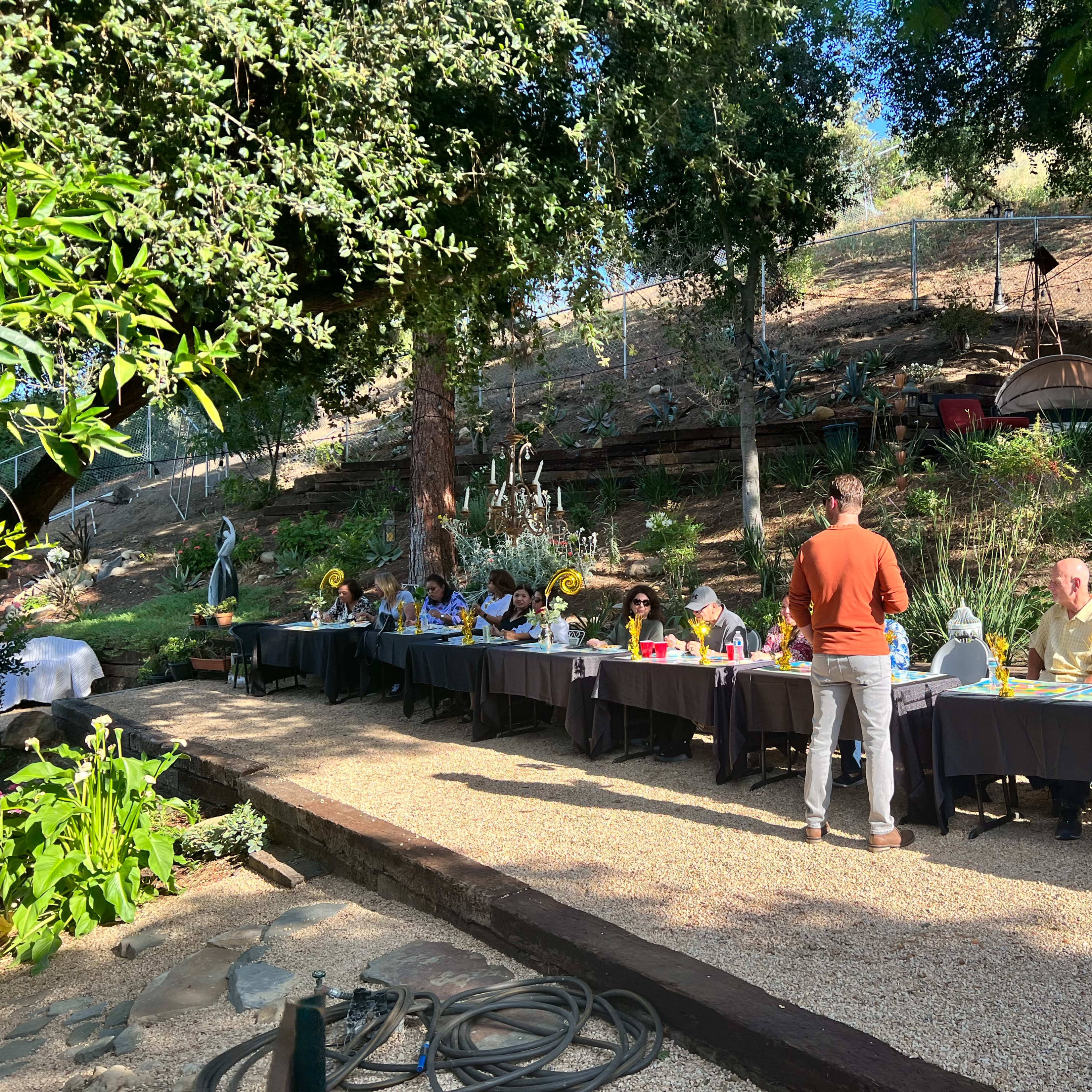A group of people sits at long tables set up outdoors in a garden-like area surrounded by trees and greenery.