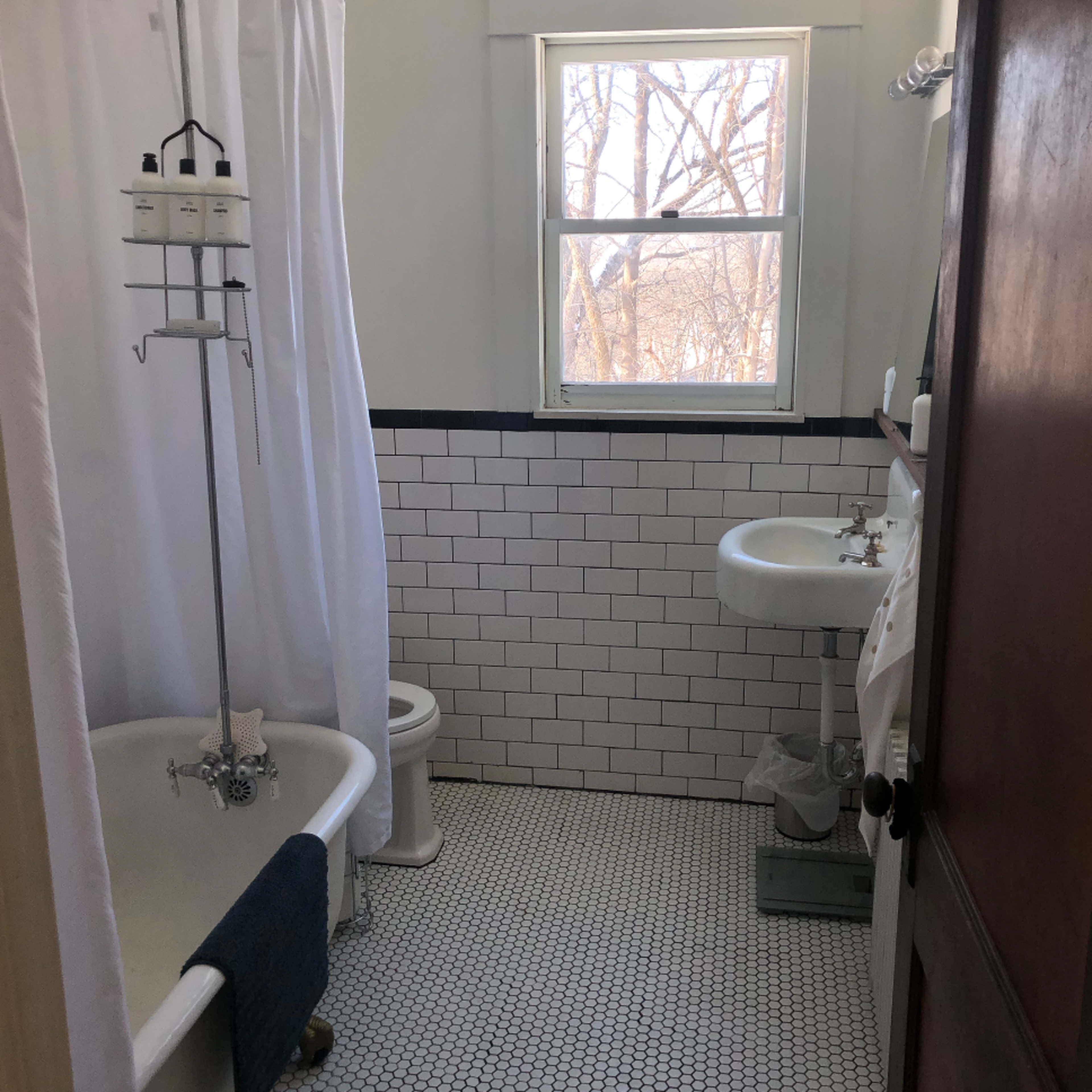 A bathroom featuring a clawfoot bathtub, a white shower curtain, a sink, a toilet, and a window with a view of bare trees outside.