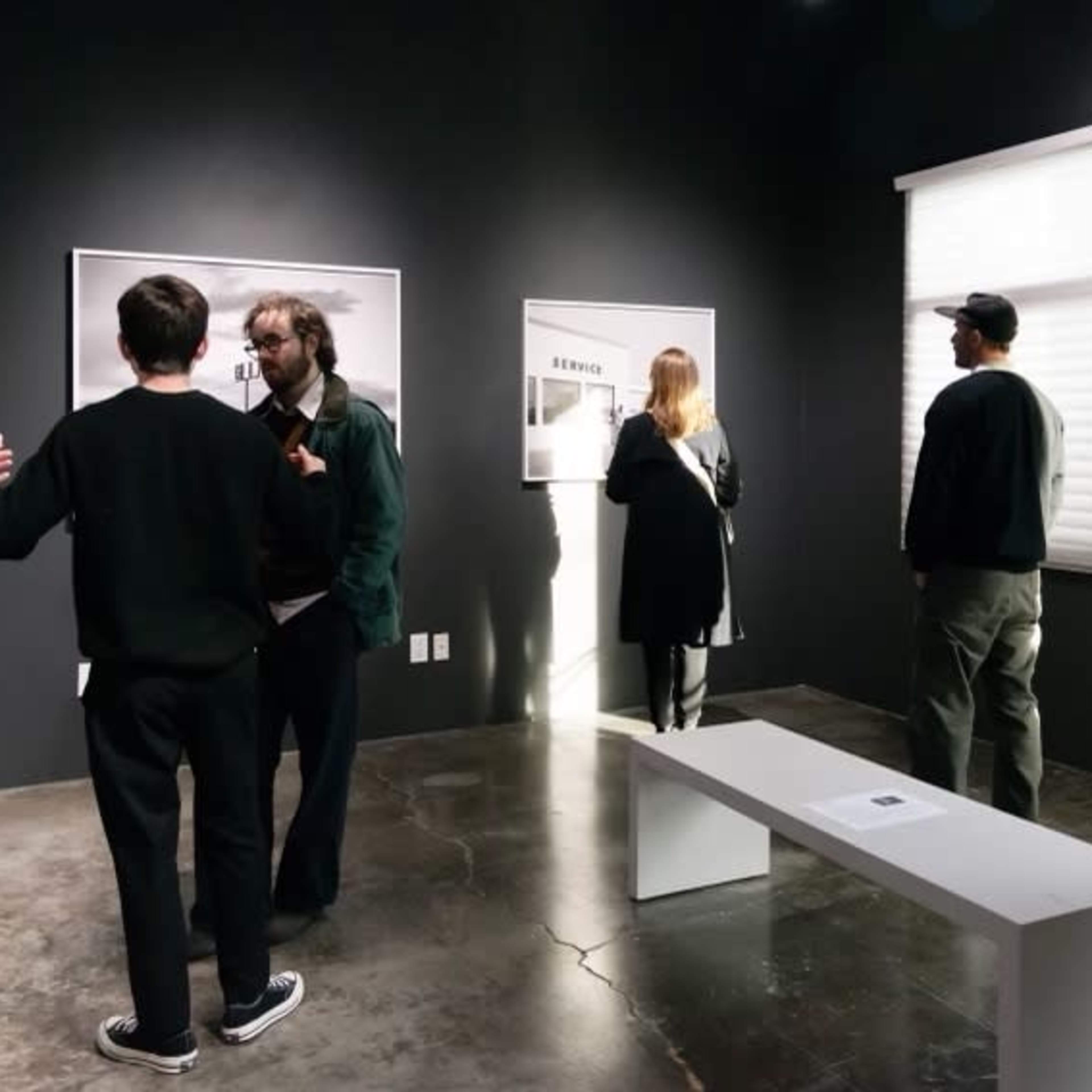 A group of people observes artwork in a dimly lit gallery with black walls and polished concrete floors.