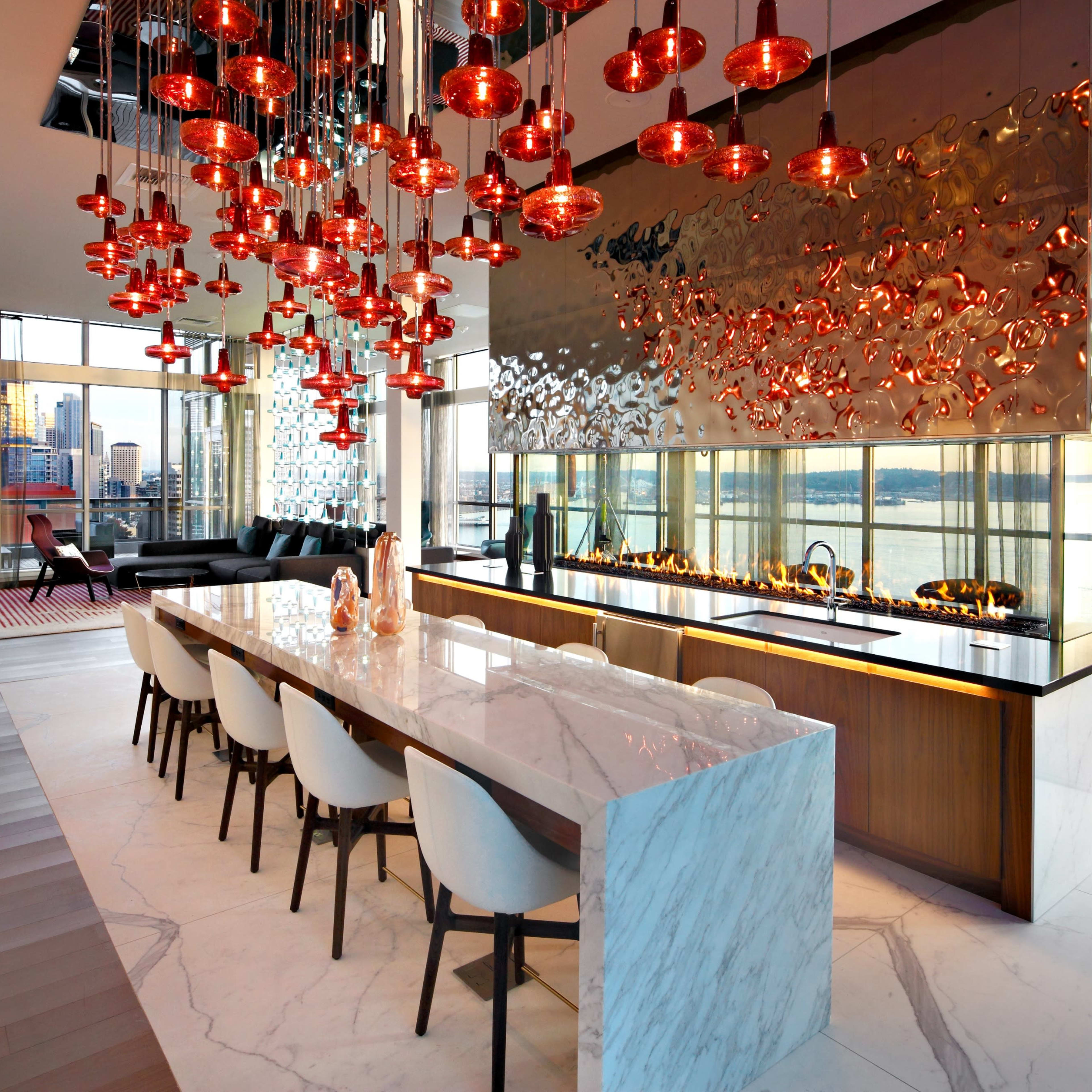 A modern kitchen features a long marble island with seating, surrounded by stylish pendant lights and large windows showcasing a city view.