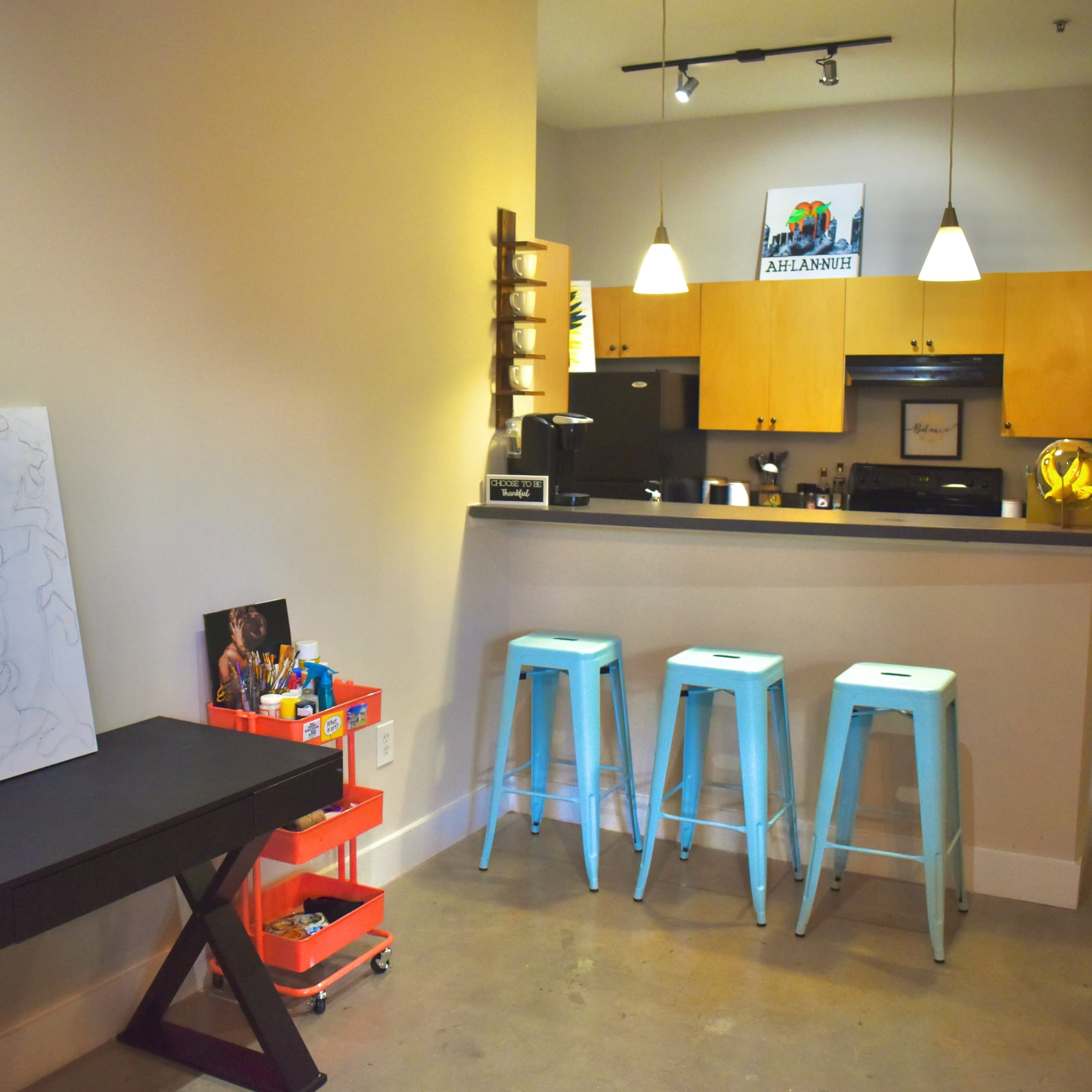 A modern kitchen area features a bar with three light blue stools and a dark table with an art canvas nearby.