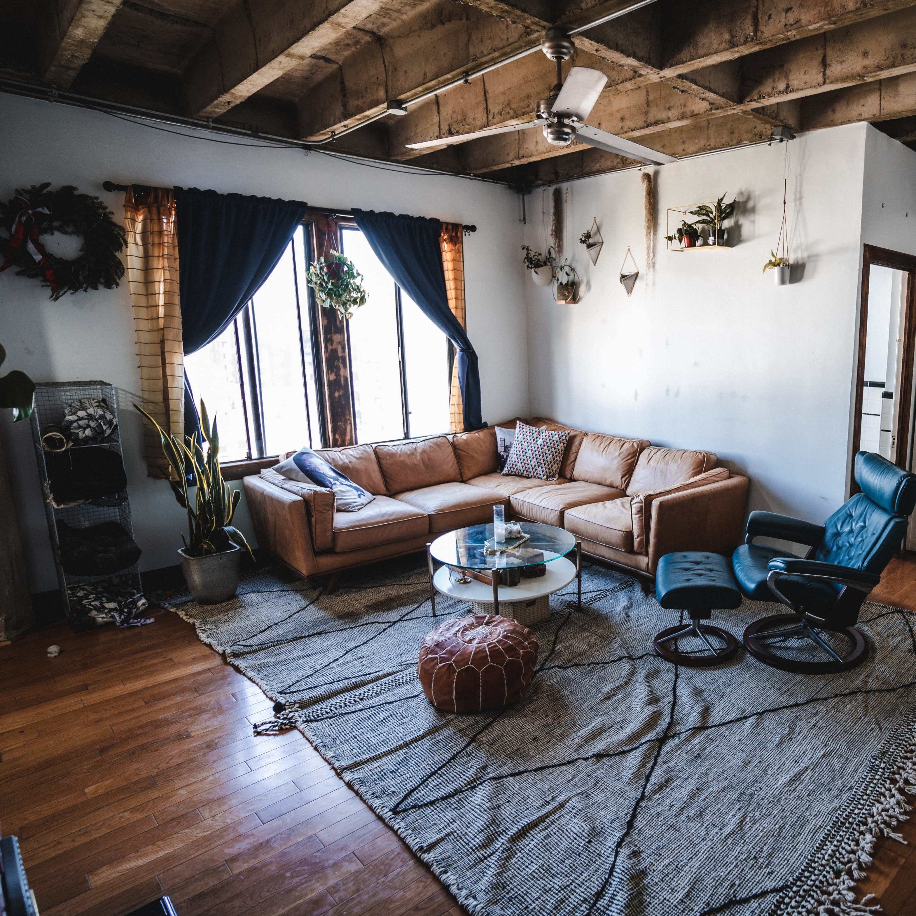 A cozy living room with a brown sectional couch, a round glass coffee table, a blue lounge chair, and a patterned area rug on hardwood flooring.