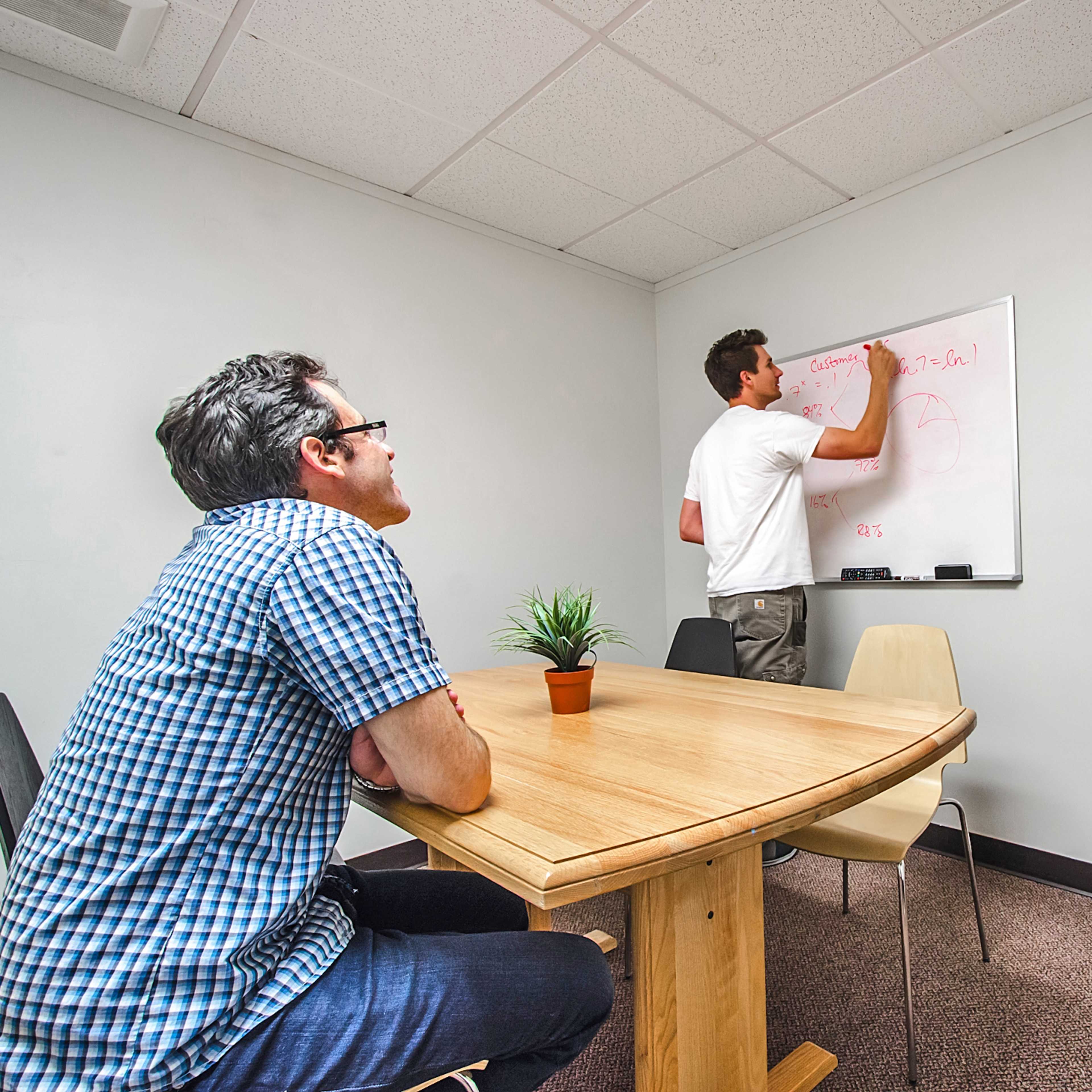 A man writes on a whiteboard while another man observes, seated at a wooden table in a small office meeting room.