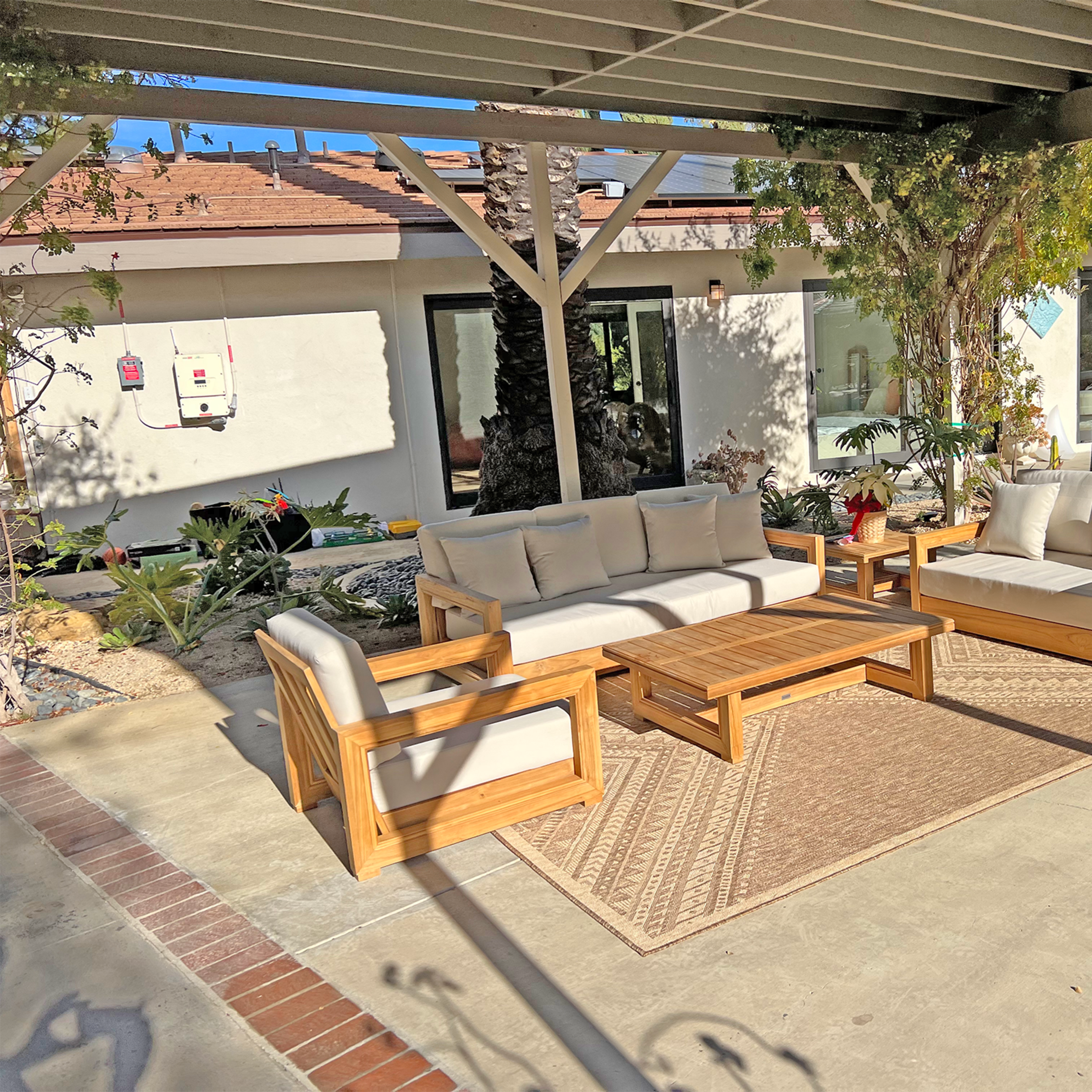A modern outdoor seating area features a wooden sofa set and coffee table on a concrete patio, surrounded by plants and adjacent to a house.