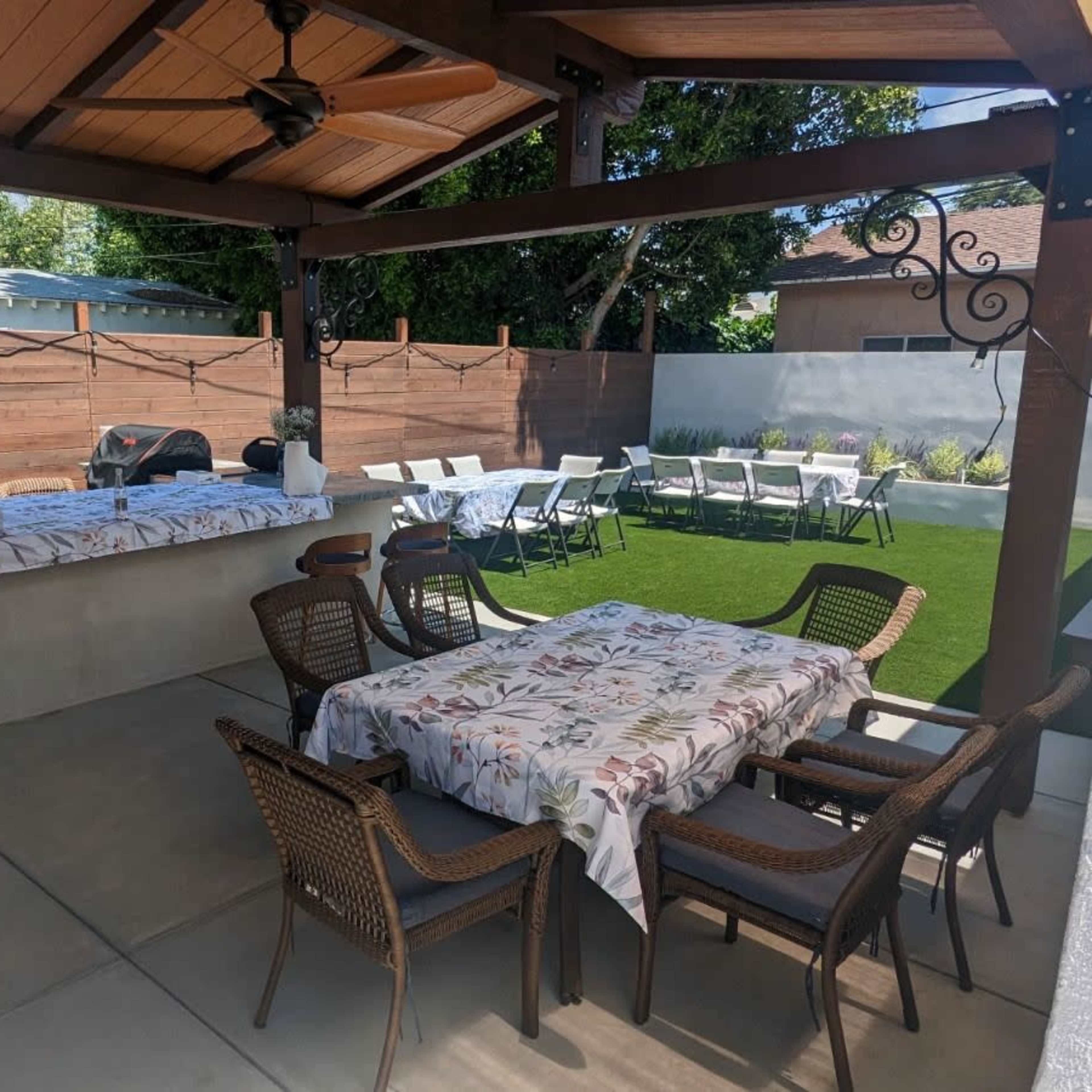 A covered outdoor patio features a table with floral tablecloths, wicker chairs, and an adjacent area with additional seating and a barbecue setup.