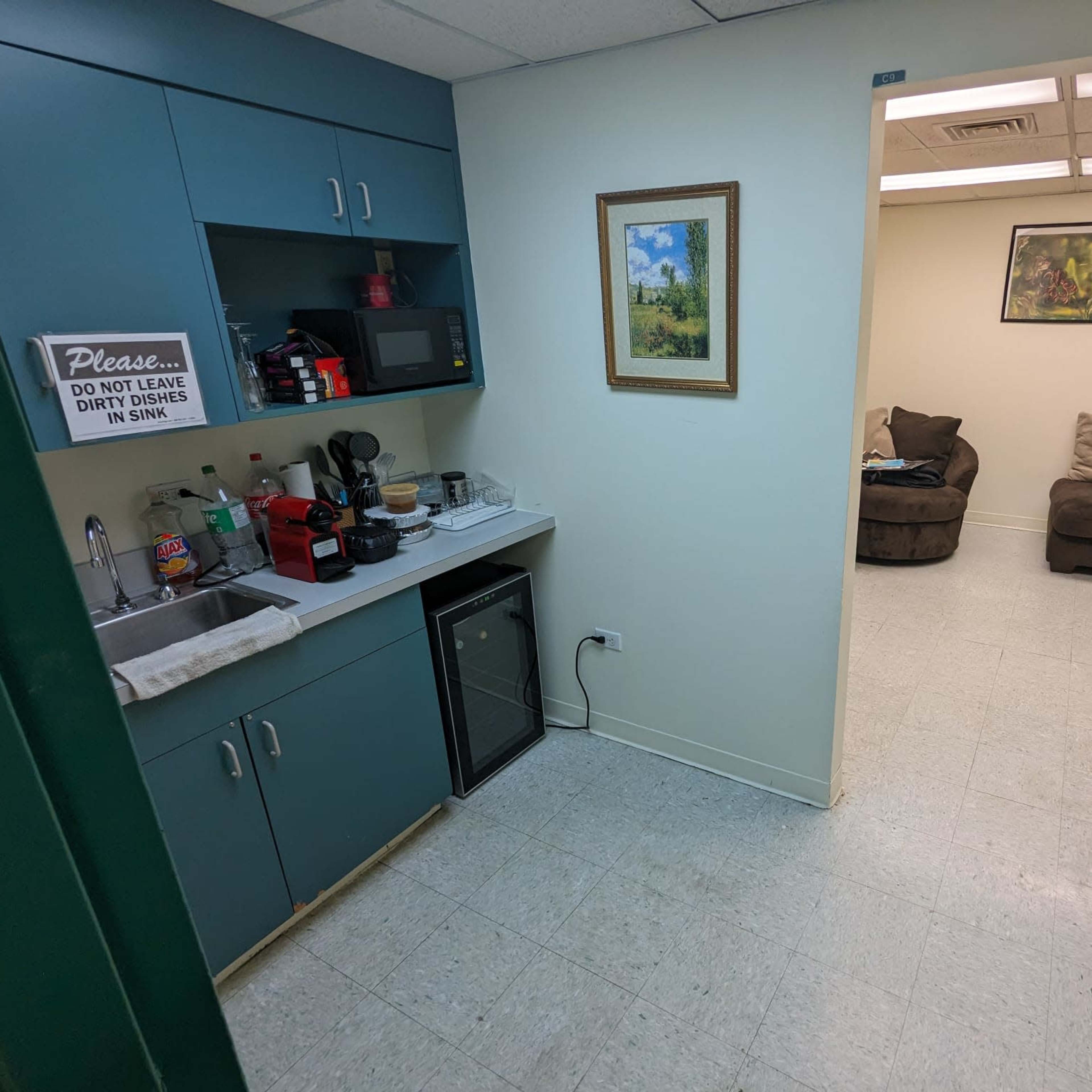A small kitchenette with blue cabinets, a microwave, and a coffee maker, adjacent to a room featuring two brown chairs.