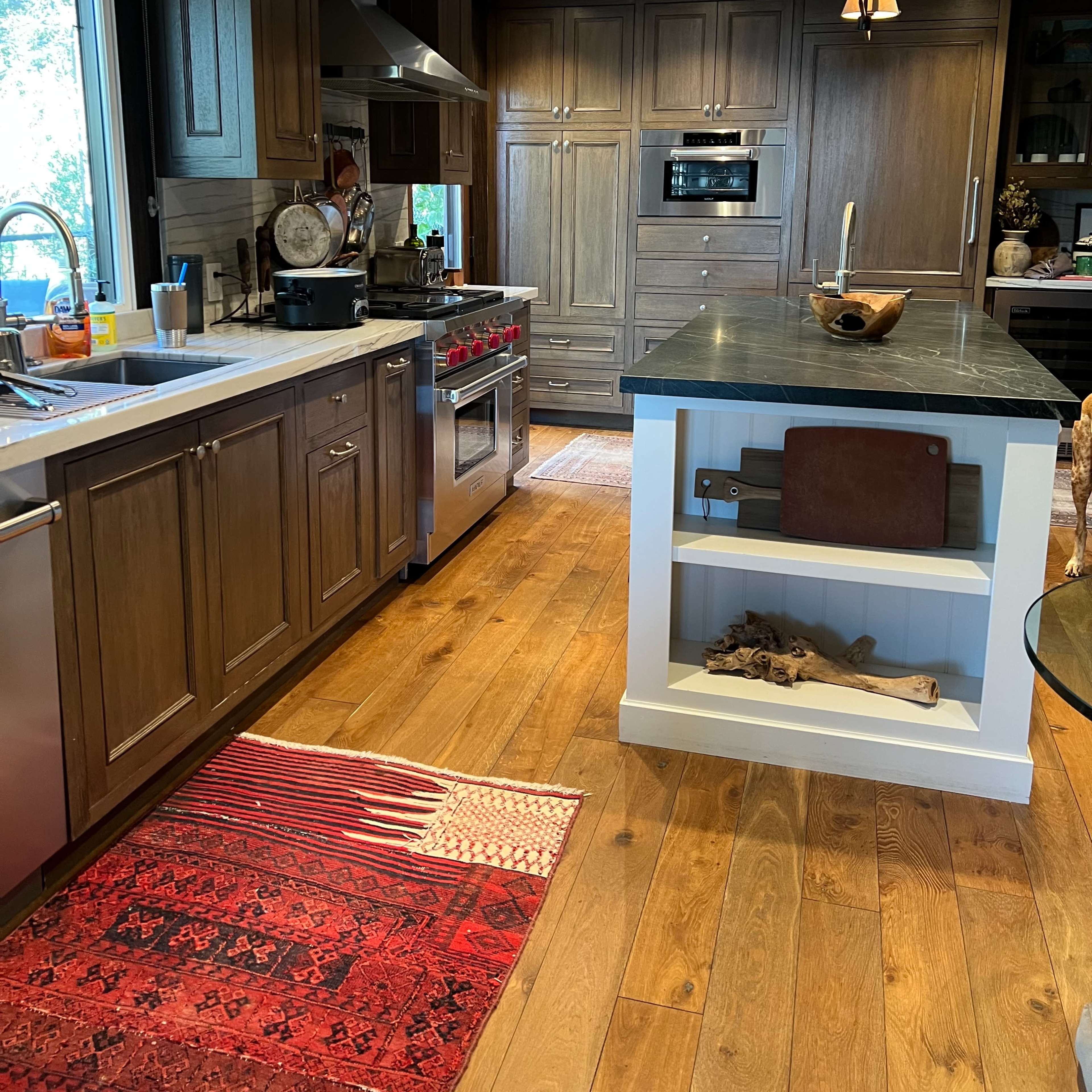 The kitchen features wooden cabinets, stainless steel appliances, a central island with a white base, and a red area rug on a hardwood floor.