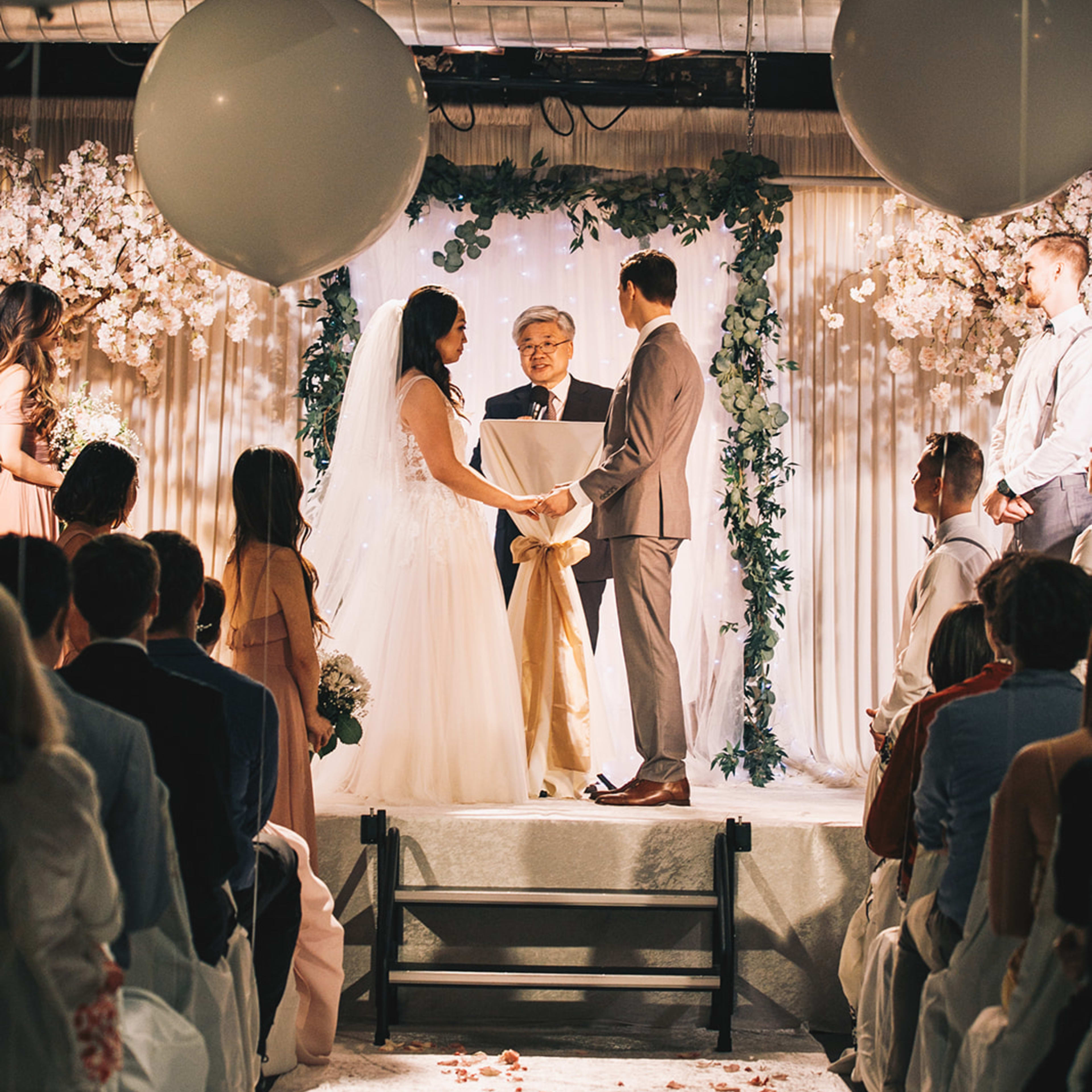 A couple stands at an altar decorated with flowers, exchanging vows in front of an officiant, while guests seated on either side watch.