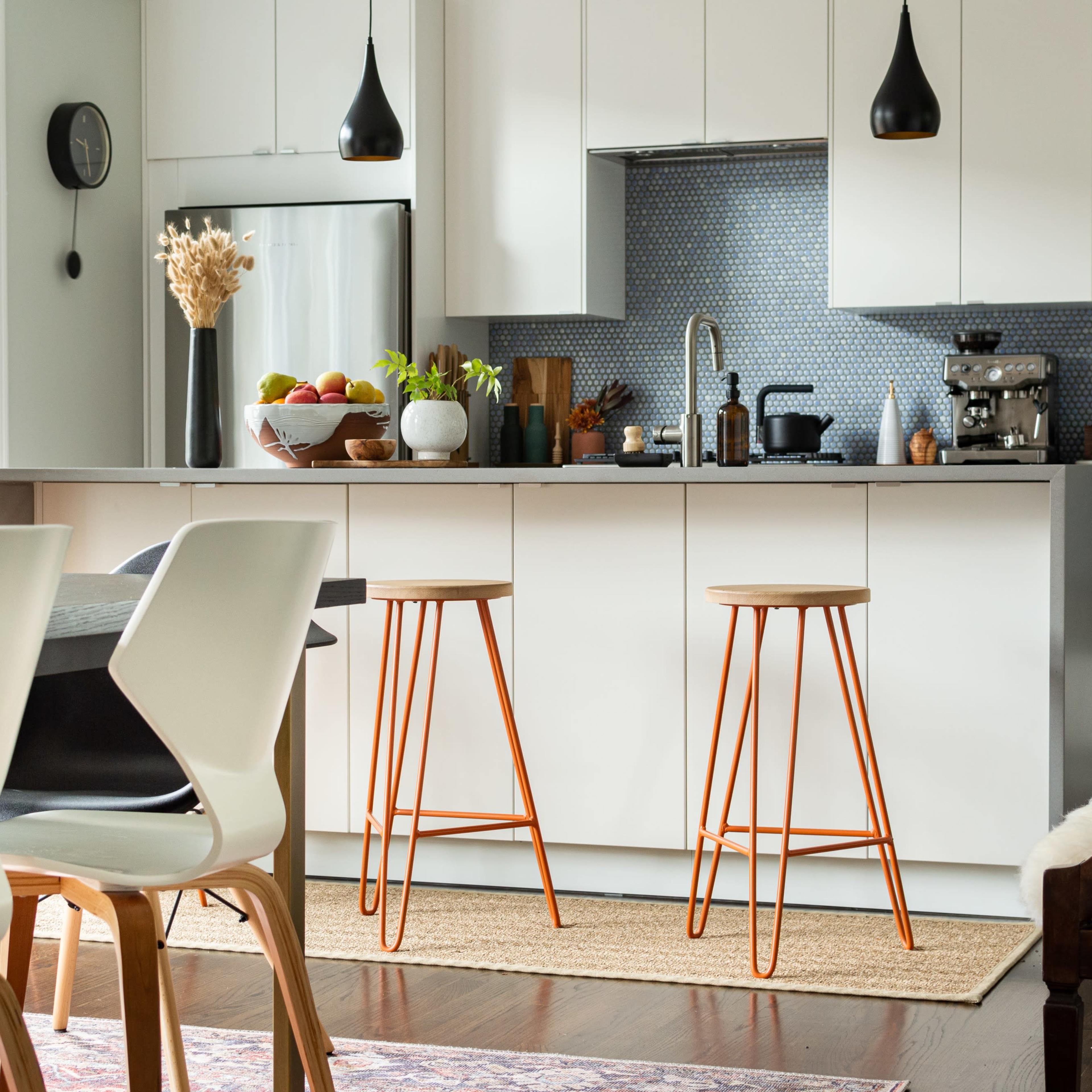 A modern kitchen features white cabinetry, a blue tiled backsplash, two metal bar stools with wooden tops, and a dining area with white chairs.