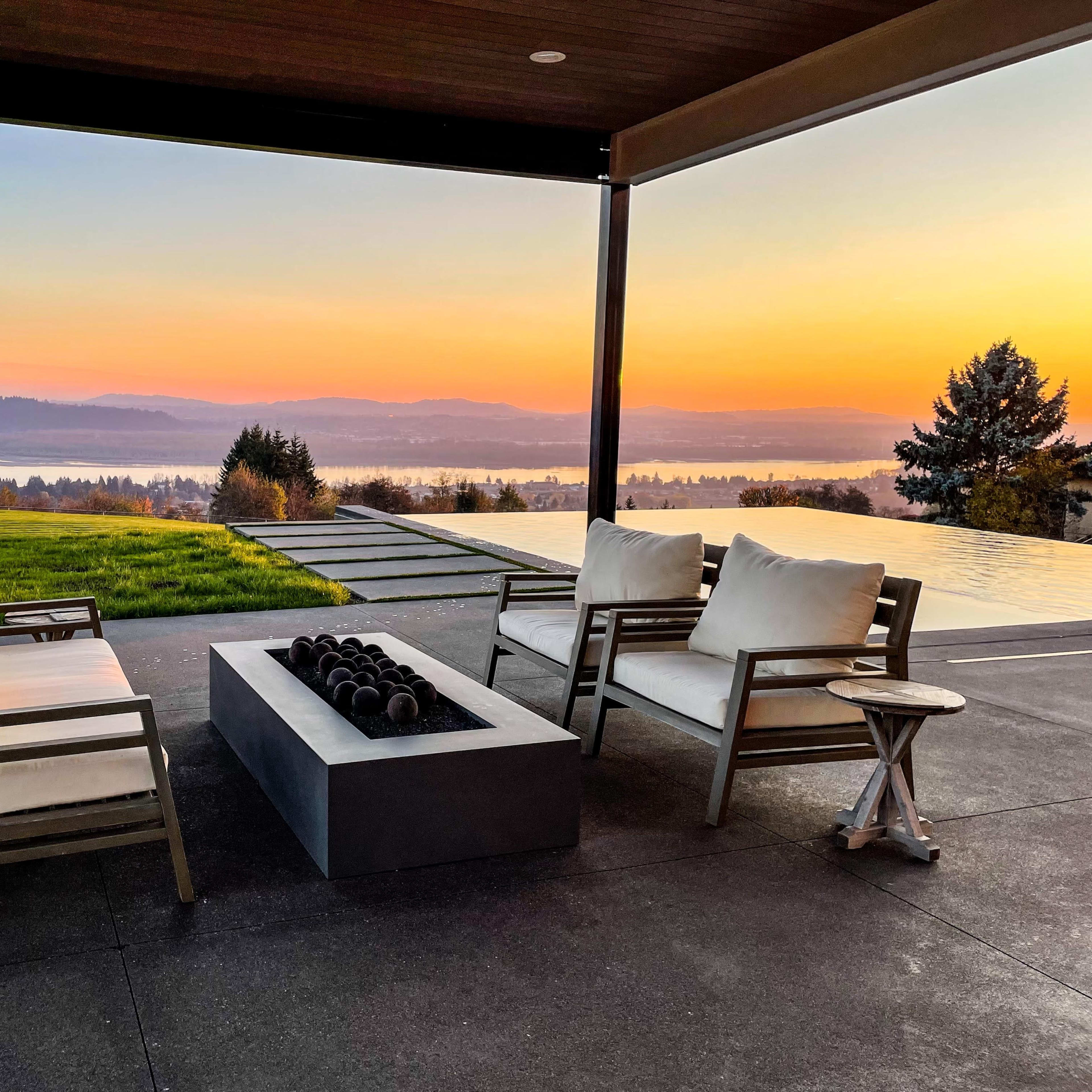 A patio area features two chairs and a fire pit overlooking a lake at sunset.