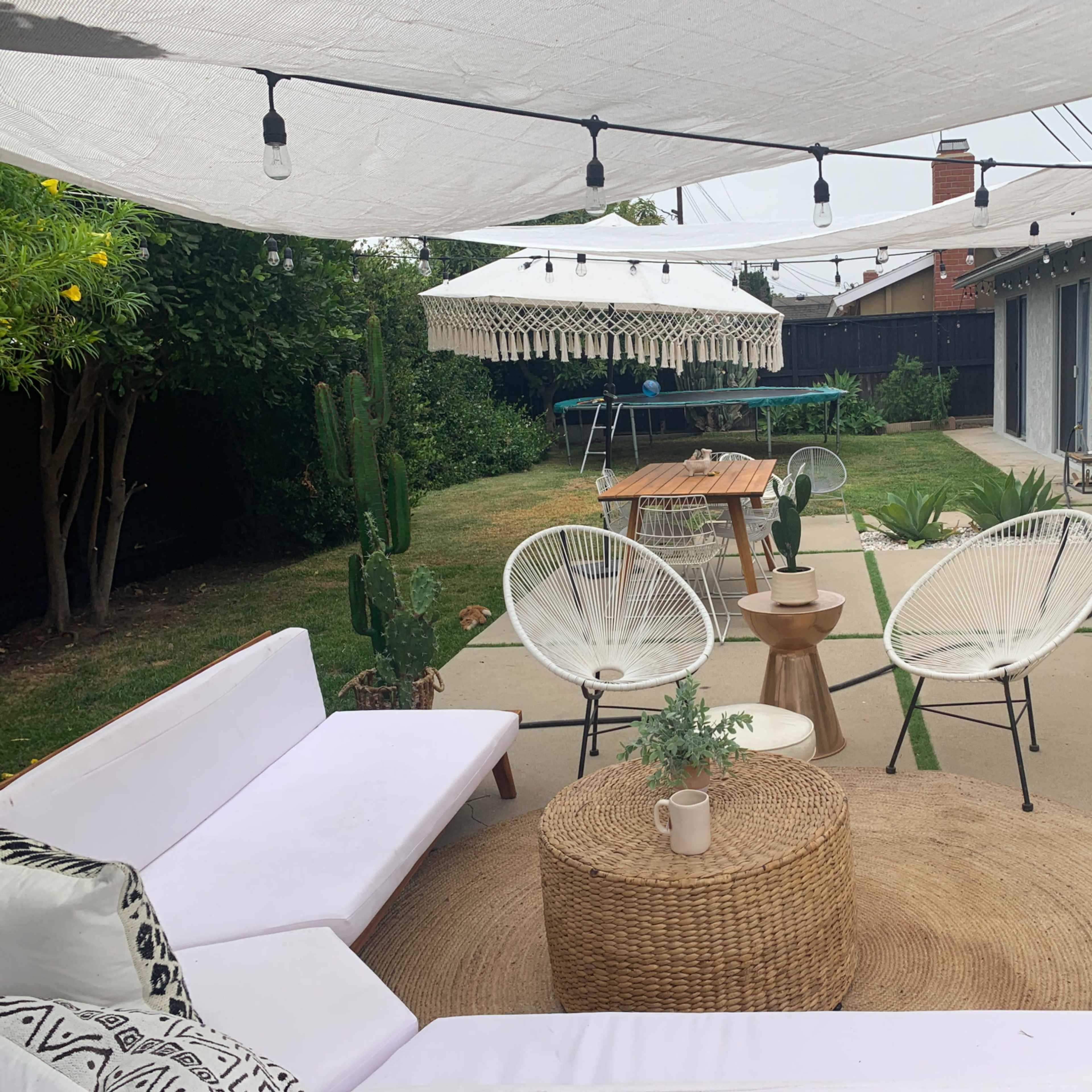 A backyard patio features a seating area with white chairs, a round table, and string lights overhead, surrounded by greenery and potted plants.