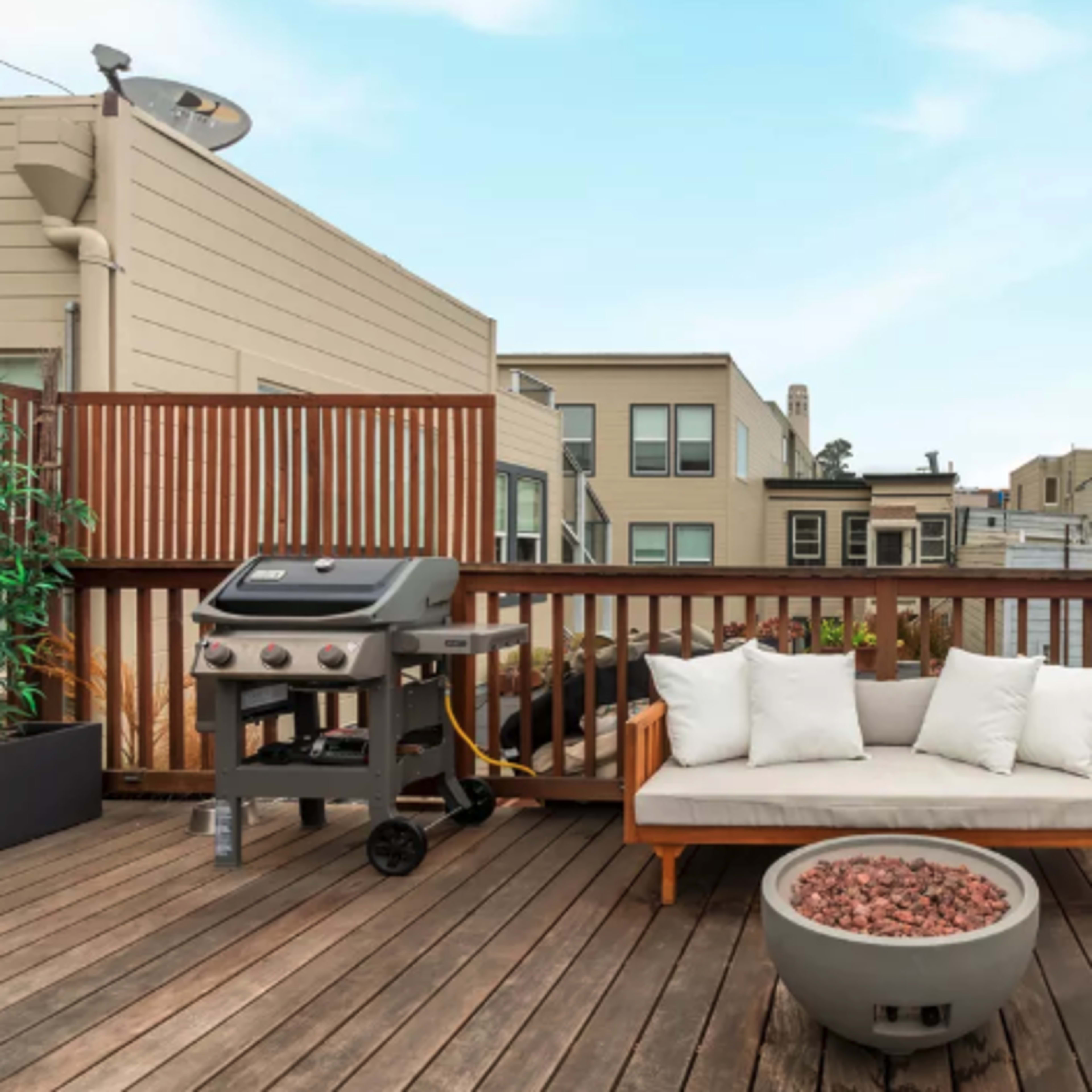 A wooden deck with a sofa, a fire pit filled with stones, and a grill, surrounded by buildings in the background.