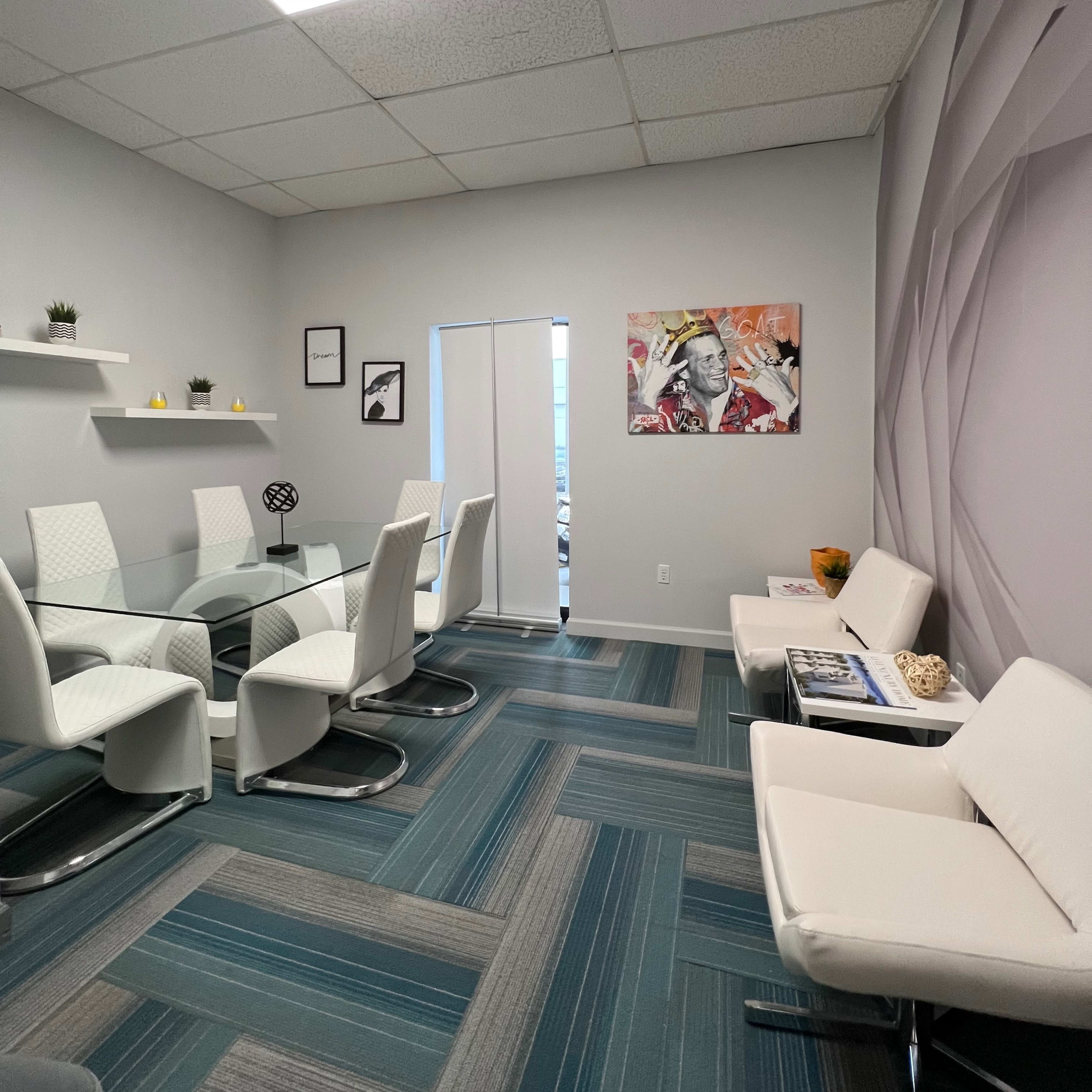 A modern office space with a glass meeting table surrounded by white chairs, light gray walls, and a colorful artwork hanging on one side.