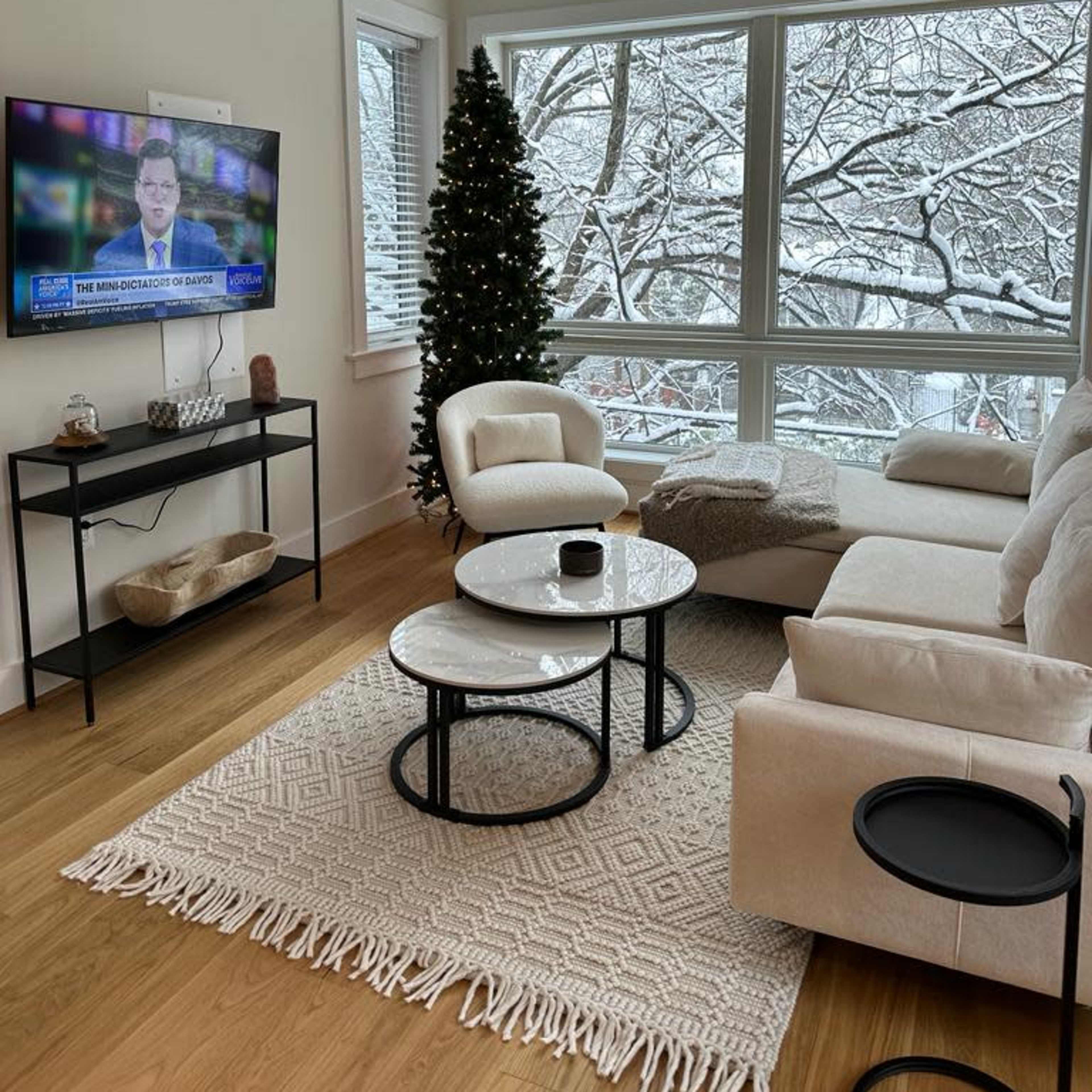 A modern living room with a television displaying news, a Christmas tree, and large windows overlooking a snowy landscape.